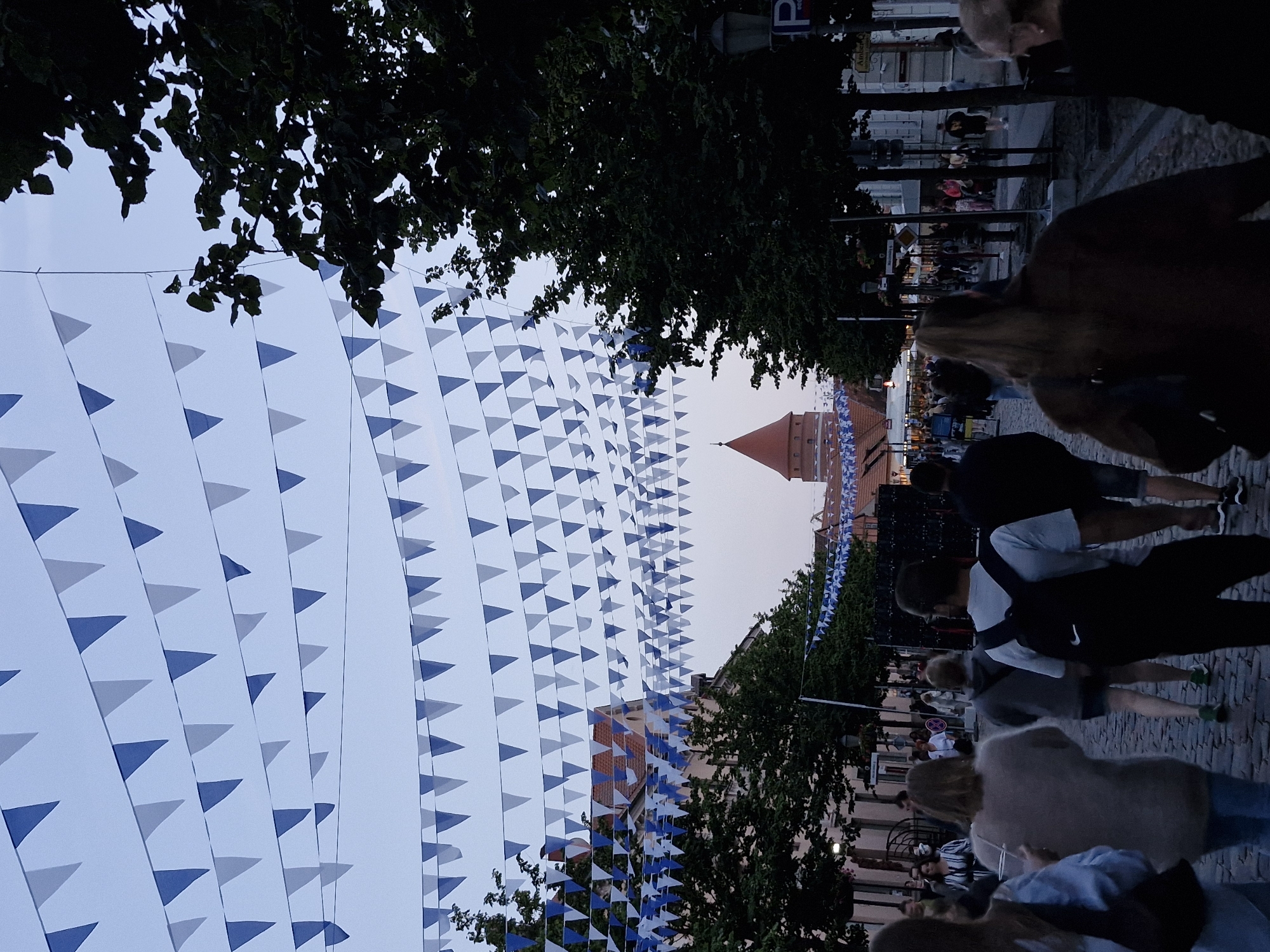 The image depicts a street scene in Klaipėda, Lithuania, likely during an evening event. Numerous triangular flags in blue and white are strung across the street, creating a festive atmosphere. The street is paved with cobblestones and lined with trees and buildings. In the background, a distinctive red-roofed tower is visible. Several people are walking along the street, some in small groups, suggesting a gathering or festival. The lighting indicates it is dusk or early evening. The scene captures a lively urban environment with decorative elements.