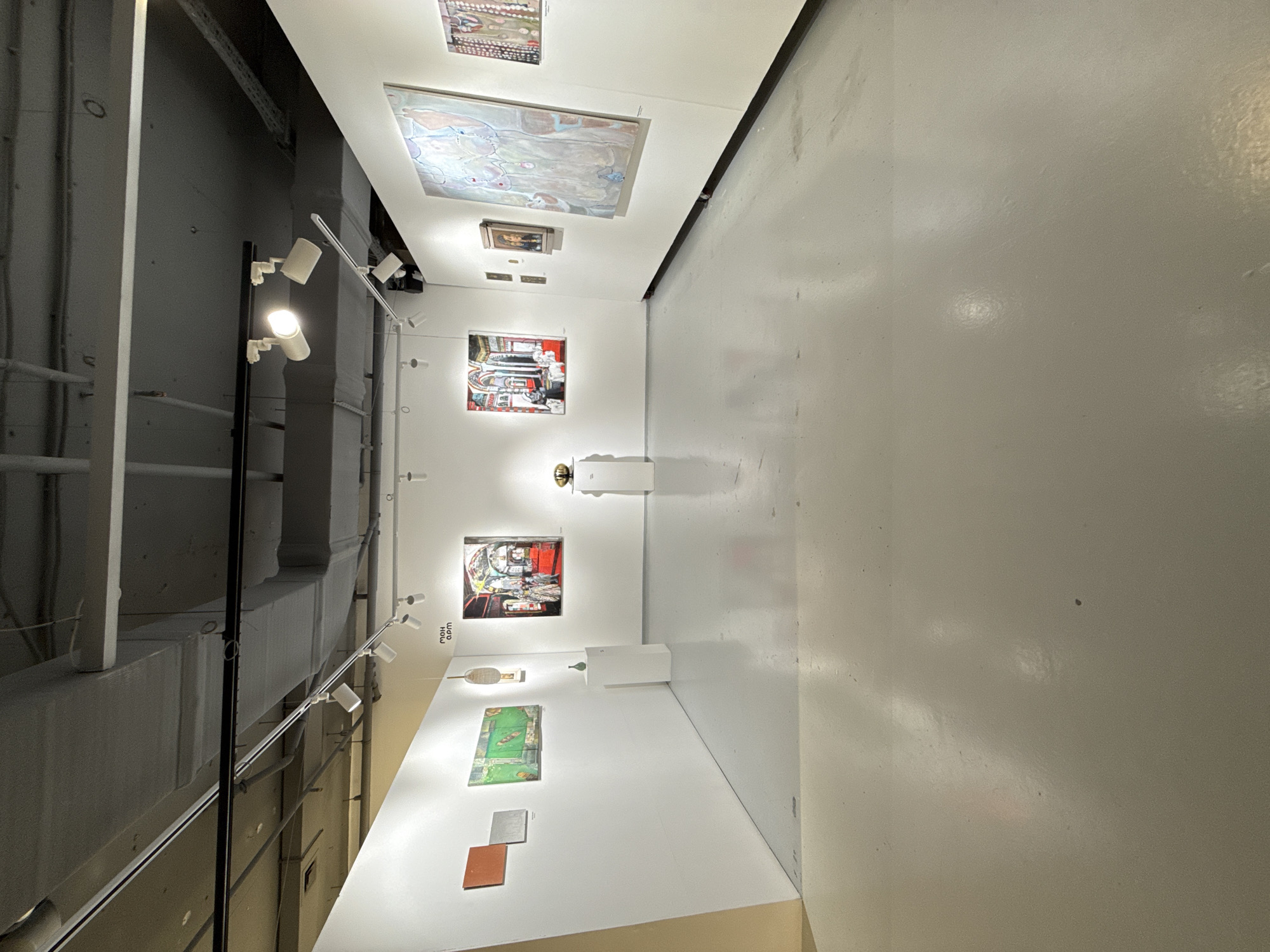 The image shows an art gallery in Unknown, Unknown. There are various paintings hanging on the white walls. A track lighting system is installed on the left side of the wall, and the floor is white. No people are visible in the image.