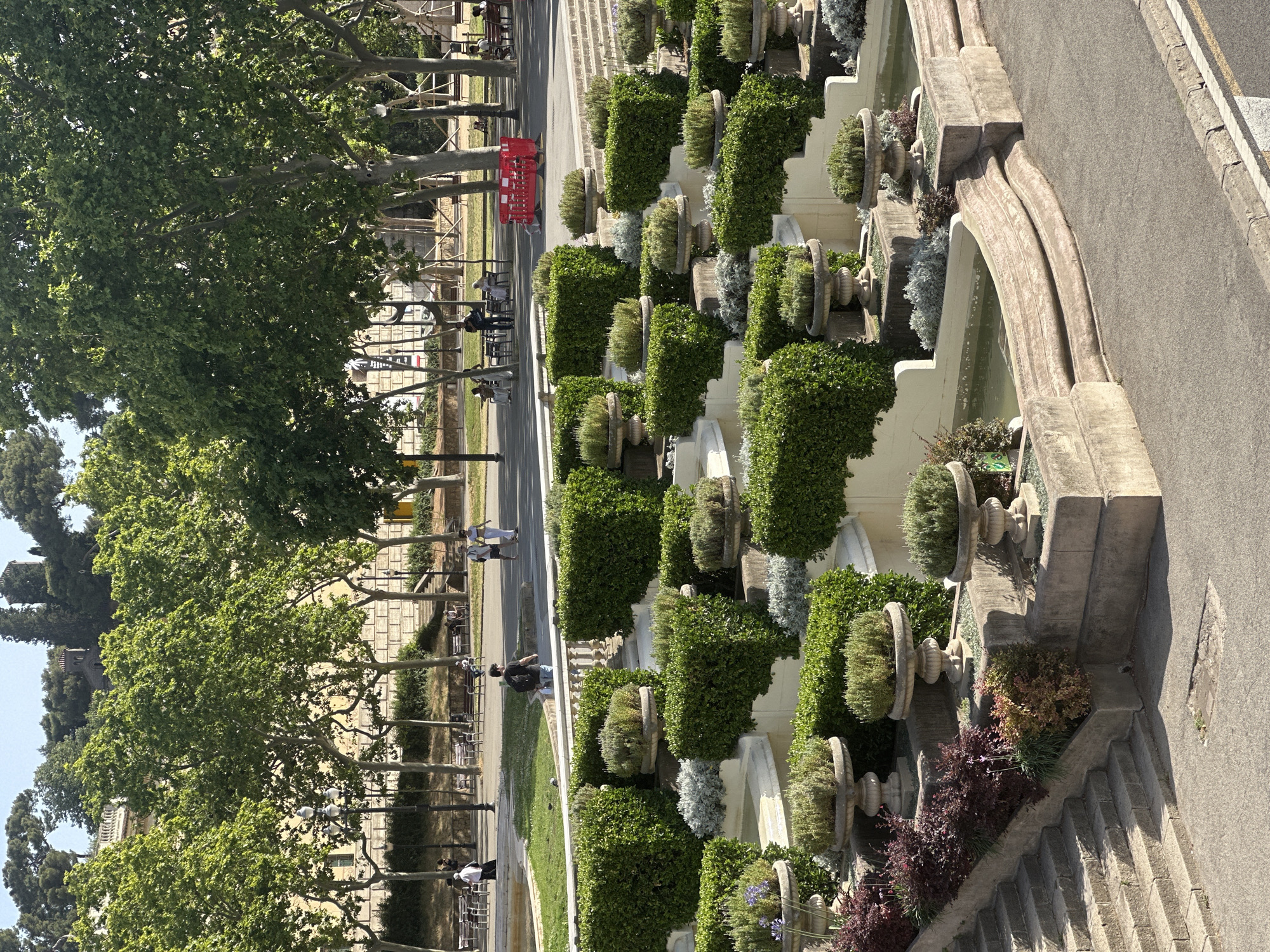 The image depicts a tiered garden landscape with geometric topiaries and ornate planters filled with greenery. The garden is situated on a slope, with stone steps leading up from the lower right of the frame. In the background, several people are visible walking and sitting on benches in a park area, with mature trees providing shade. A building with a classical facade is partially visible behind the trees on the left. The scene appears to be bathed in bright sunlight. This landscape is characteristic of Park Güell in Barcelona, Spain.