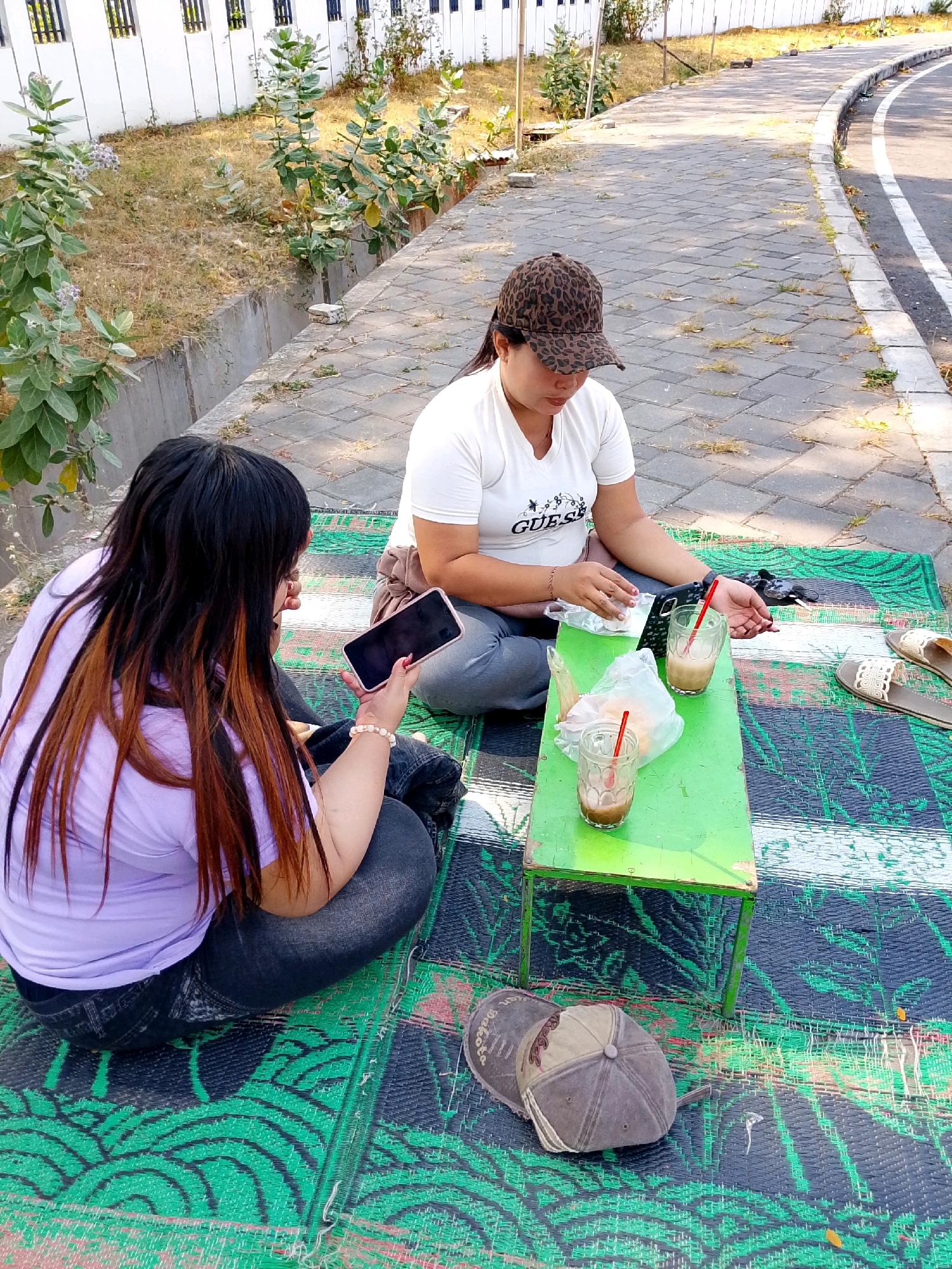 Two women are sitting on a green and black mat on a paved area next to a road in Bulusari, Indonesia. The woman on the left has long, dark hair with reddish-brown highlights and is wearing a lavender t-shirt and dark jeans. She is holding a smartphone in her right hand and looking at it. A beaded bracelet is on her left wrist. The woman on the right is wearing a white t-shirt with the word "GUESS" printed on it and a leopard-print baseball cap. She is looking down at her hands, which are near a small, green table. On the table are two glasses with drinks and red straws, and two white packages, possibly food. To the right of the table, a pair of sandals are placed on the mat. Two baseball caps are on the mat near the table. The paved area appears to be a walkway or sidewalk, and a road with a white line marking is to the right of the walkway. In the background, there is a white fence and some dry vegetation.