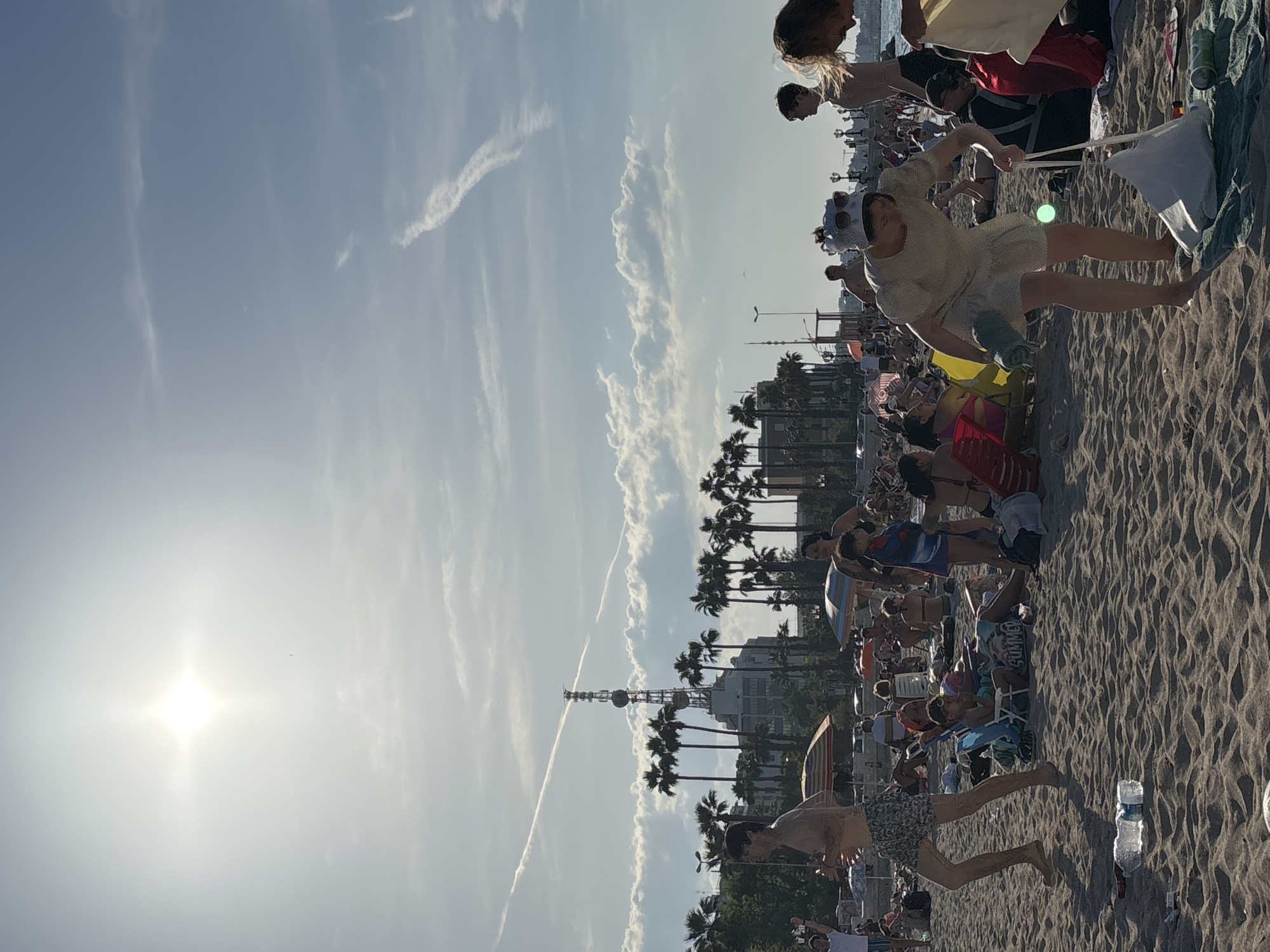 This image captures a vibrant and crowded beach scene on a sunny day. The sky is a clear blue with scattered white clouds and a few faint contrails, suggesting good weather. The sun is a prominent, bright light source in the upper left of the frame, casting long shadows on the sand and indicating it is likely mid-morning to early afternoon.
Numerous people are visible across the sandy beach. In the foreground, a person wearing swim shorts walks barefoot on the rippled sand, next to a discarded plastic water bottle. Slightly to their right and further back, another individual, dressed in light-colored clothing and sunglasses, stands holding a white flag or a kite with strings.
Further into the mid-ground and background, many other beachgoers are seen relaxing, some sitting on beach chairs under colorful umbrellas, while others are on towels. The shoreline is lined with a row of tall palm trees, beyond which urban buildings are visible, suggesting a coastal city or resort area. A tall, distinctive transmission tower or antenna stands out against the sky in the distance, serving as a notable landmark.
The overall scene conveys a cheerful and leisurely atmosphere, with people enjoying the warmth and sunshine characteristic of a summer beach day. No discernible text is clearly visible in the image.