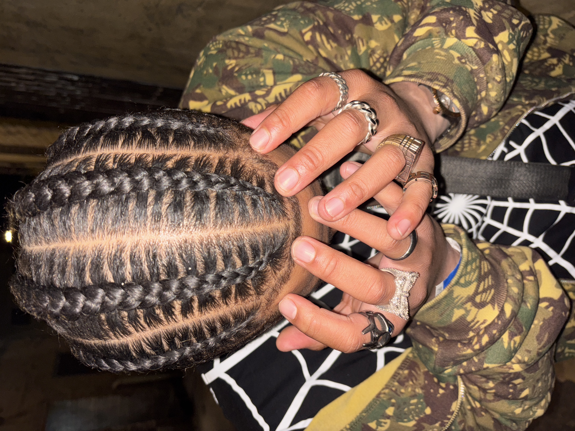 A close-up shot shows a person with cornrow braids, their head angled to the left. The braids are neat and tight against the scalp. Multiple rings adorn the person's fingers, including silver and gold bands with varying designs. The person is wearing a camouflage jacket and a black and white patterned garment underneath. The background is dark and out of focus. The location is not discernible from the image provided.