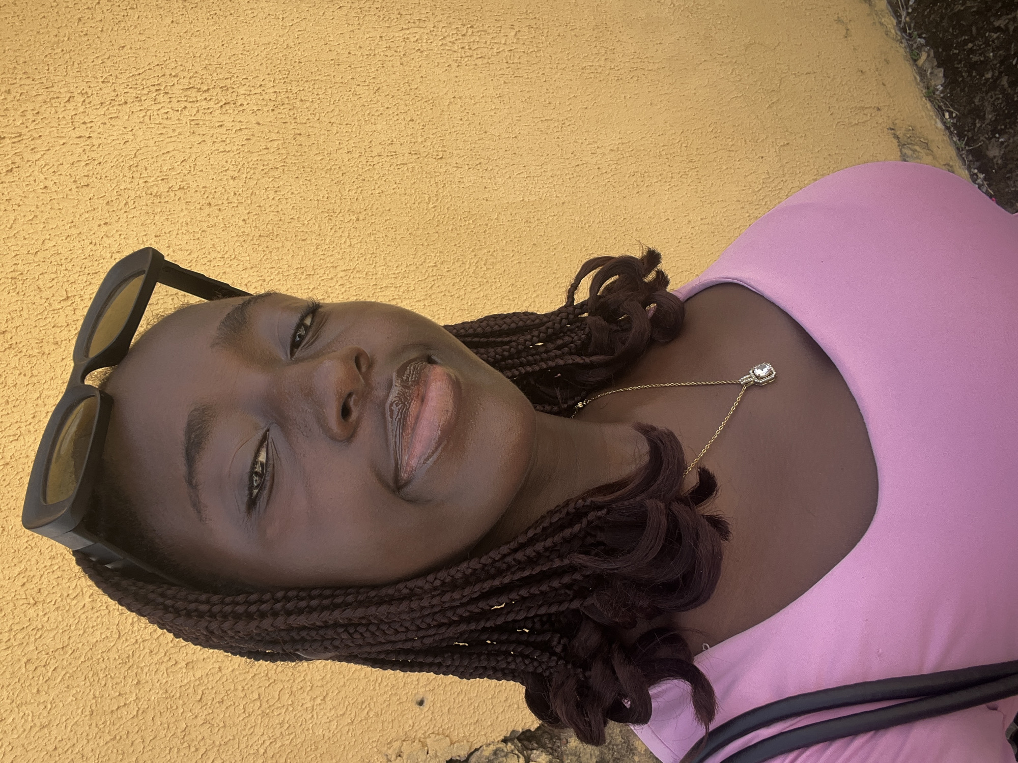 The image shows a young Black woman with brown braided hair, wearing a pink top and gold necklace. Sunglasses are perched on top of her head. She is positioned against a textured yellow wall, which appears to be an exterior. The location is suggested to be in Abuja, Nigeria, based on the prompt. There is a dark-colored object near her body; it appears to be a strap or handle. The angle of the image suggests the photo was taken from a slightly elevated perspective.