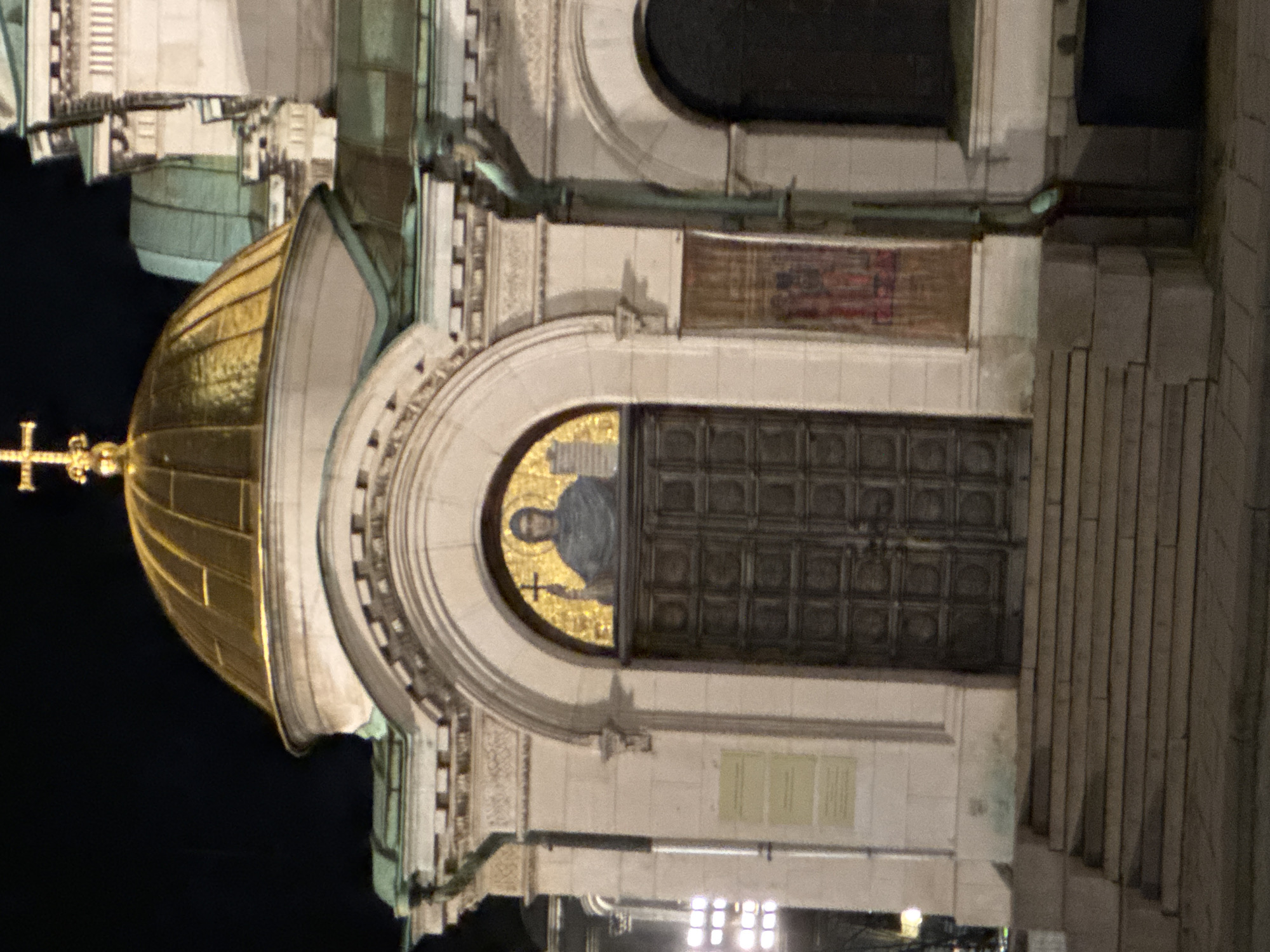 The image depicts a portion of the Alexander Nevsky Cathedral in Sofia, Bulgaria, at night. Visible are a golden dome with a cross on top, an arched entrance above a large, dark door, and adjacent steps. The archway contains a mosaic of a person, possibly a religious figure. Architectural details are visible on the building's facade, including decorative carvings and a drainpipe. The background is mostly dark, suggesting a nighttime setting. Bright light fixtures are visible at the bottom of the image. The building appears to be constructed from a light-colored stone.