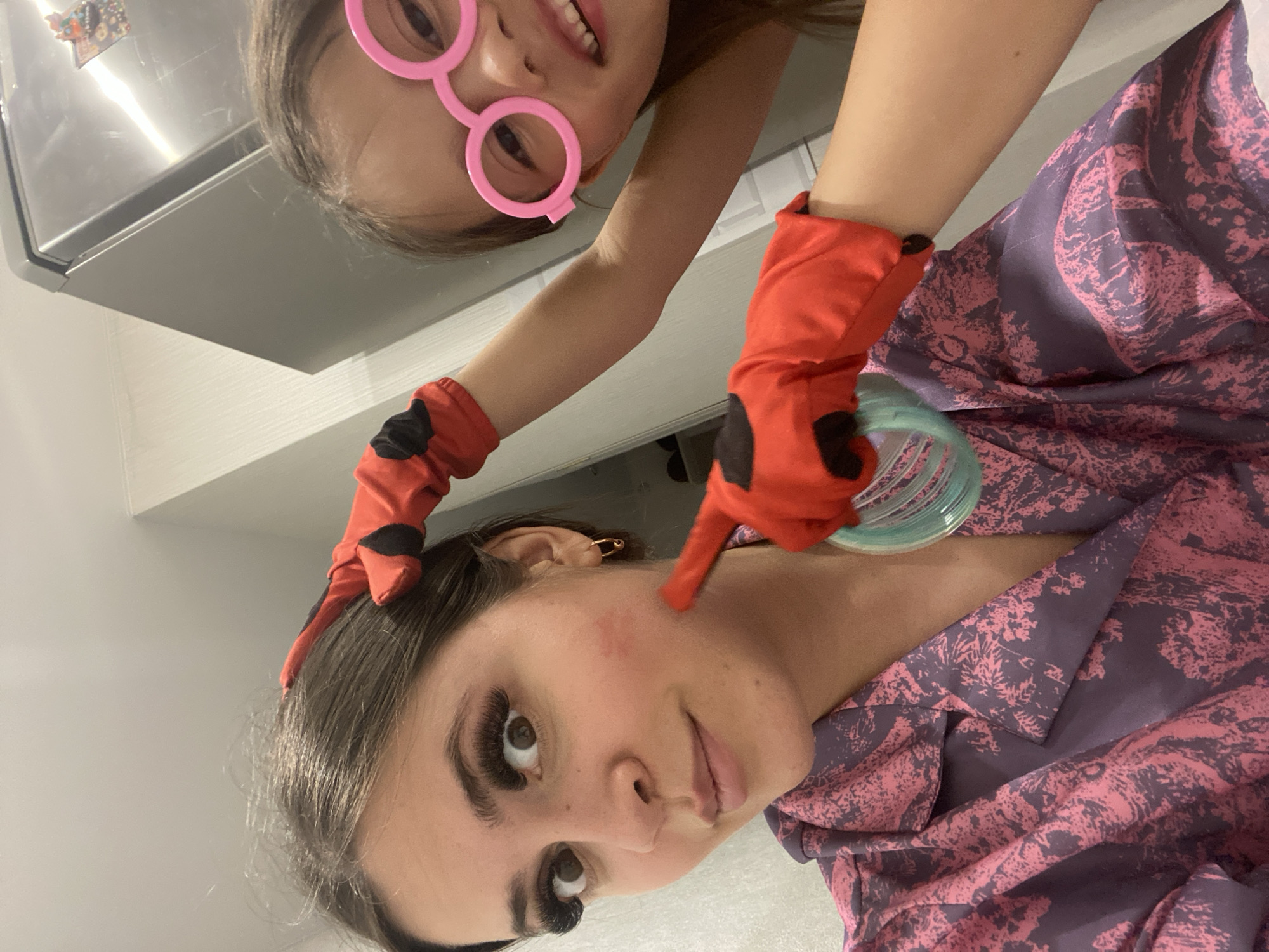 This indoor photograph captures a playful interaction between a young girl and an adult woman, possibly in Chaikovsky, Russia. The image is rotated 90 degrees clockwise, presenting the girl upside down at the top and the woman on her side looking upwards.
The young girl has brown hair and is wearing novelty pink, circular-framed glasses. She displays a wide, cheerful smile, revealing her teeth. Both her arms are adorned with red gloves featuring black spots, suggesting a costume such as Ladybug from Miraculous Ladybug. Her right hand, gloved, is playfully pointing at the woman's cheek.
The adult woman has dark brown hair and prominent, long eyelashes. She is dressed in a patterned garment, likely pajamas or a robe, with a purple base and intricate pink or red abstract designs. A golden safety pin is visible as an earring in her left ear. She looks towards the camera with a neutral to slightly amused expression, and a reddish mark is visible on her left cheek where the girl's finger points.
The background elements suggest a domestic setting. A reflective stainless steel appliance, such as a refrigerator or microwave, is visible, showing reflections of small decorations or magnets. Below it, a light-colored shelf or counter is present. A translucent light blue-green object, possibly a glass or cup, is partially visible near the woman's shoulder. The overall impression is a lighthearted and affectionate moment. No visible text is present in the image.