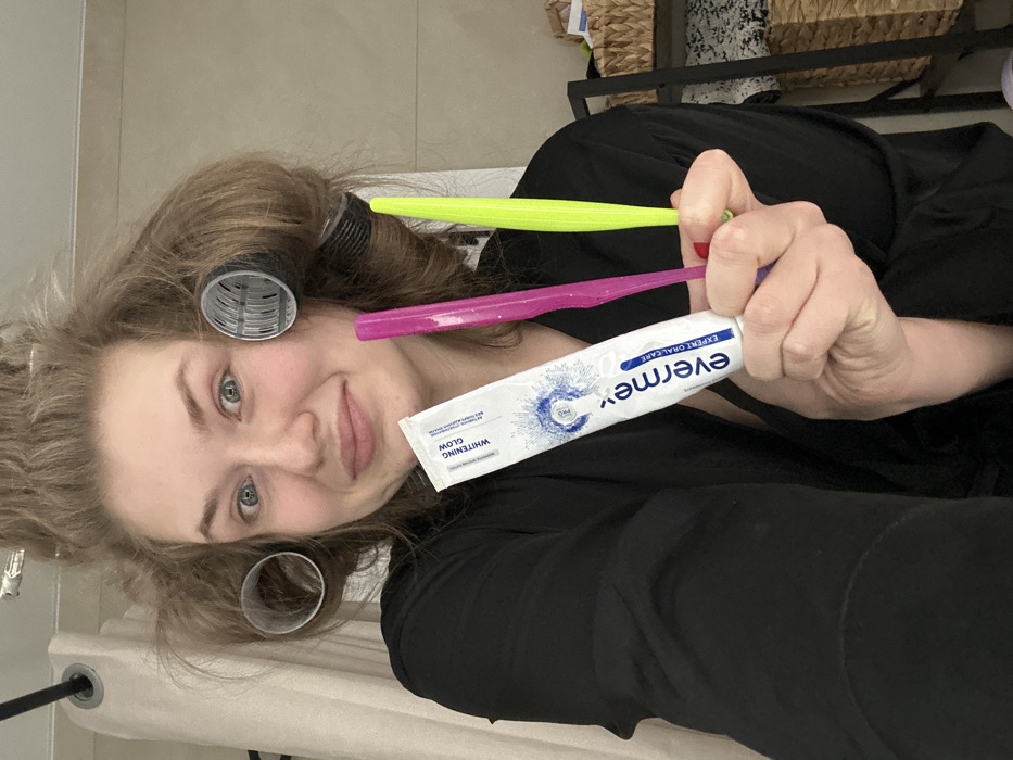 A woman with hair curlers poses with Evermex whitening toothpaste and two toothbrushes, one pink and one yellow. She is wearing a black robe and stands in a room with a shelf filled with baskets. The image is taken at an angle, showing her face and the items she holds up.