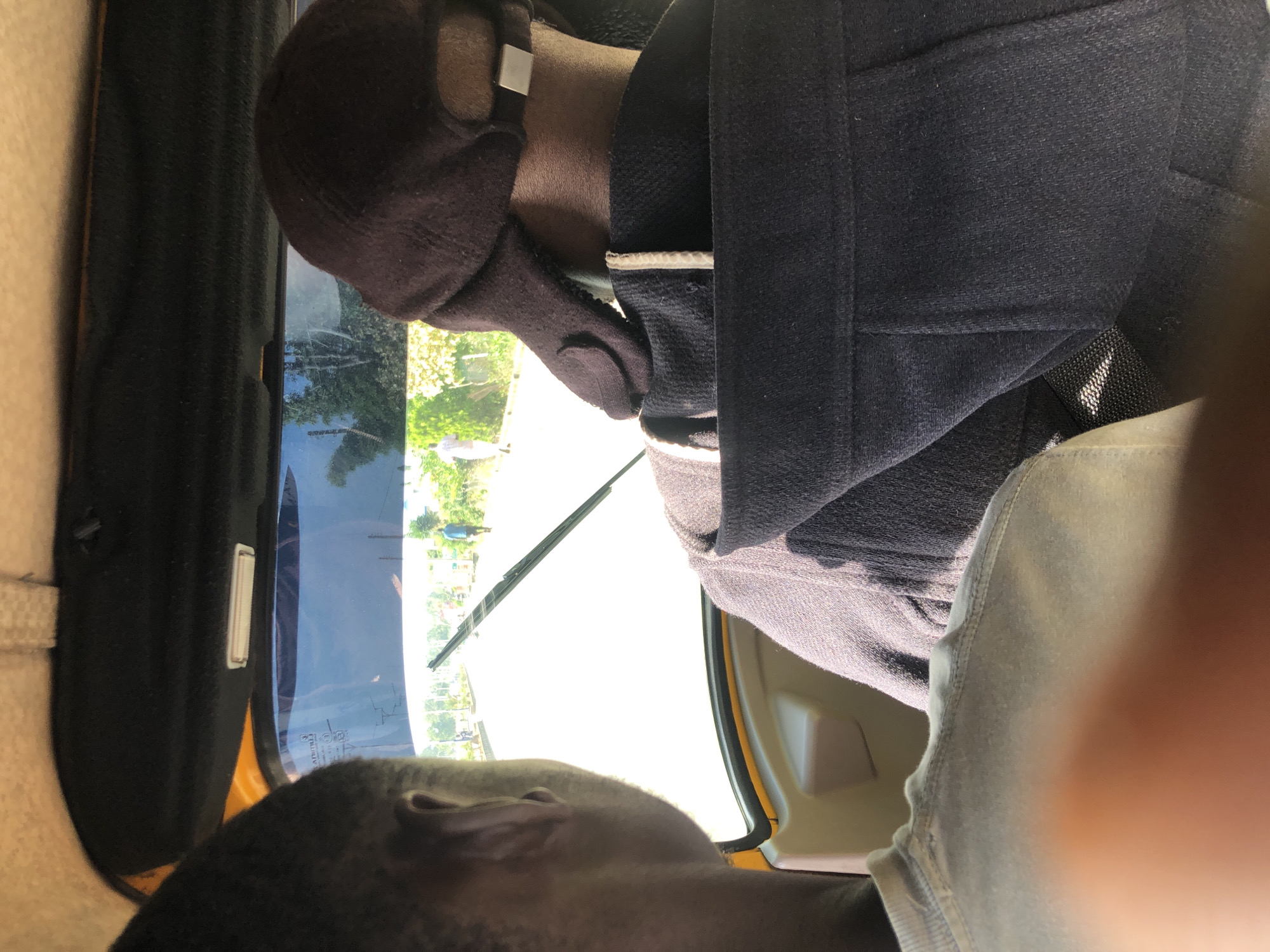 The image shows the interior of an auto-rickshaw, a common mode of transportation in Jos, Nigeria. Two individuals are visible from behind, seated inside. The person in the foreground, on the left, has short dark hair and is wearing a light-colored t-shirt. The person in the background, on the right, is wearing a dark baseball cap and a dark jacket. Through the windshield, a street scene in Jos is visible, with a few pedestrians and some trees lining the road. The sun appears to be bright, casting shadows. The overall impression is a candid snapshot of everyday life in Jos.