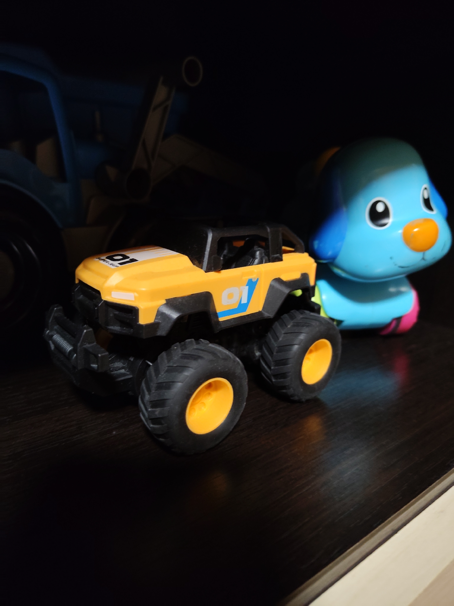 In a dimly lit indoor setting, likely a shelf or tabletop, a yellow toy monster truck with black tires and orange rims is positioned in the foreground. The truck features a "01" graphic on its hood. To the right and slightly behind the truck is a bright blue toy dog with a large orange nose and small black eyes. The background is dark and out of focus, but a hint of a larger blue toy vehicle can be seen on the far left. The environment suggests a child's play area or storage space.