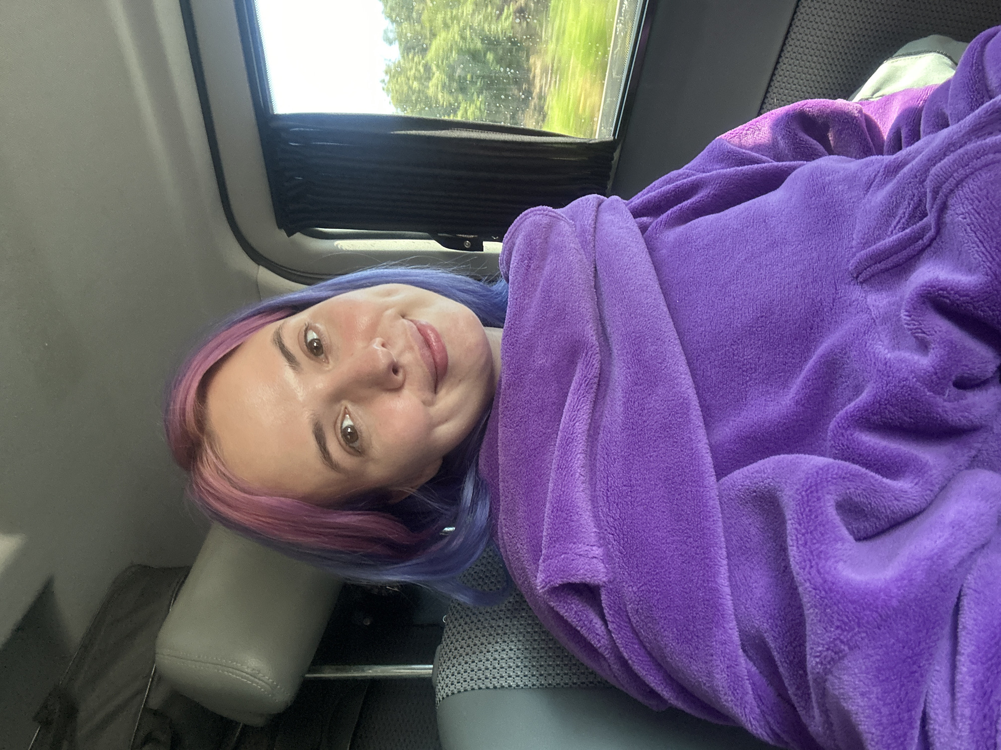 The image shows a woman with dyed pink and blue hair lying in what appears to be a vehicle. She is wearing a purple plush garment. A window is visible in the background, showing trees and greenery. The location is potentially Le Muy, France, as per the prompt's instructions, though no specific elements in the image directly confirm this.