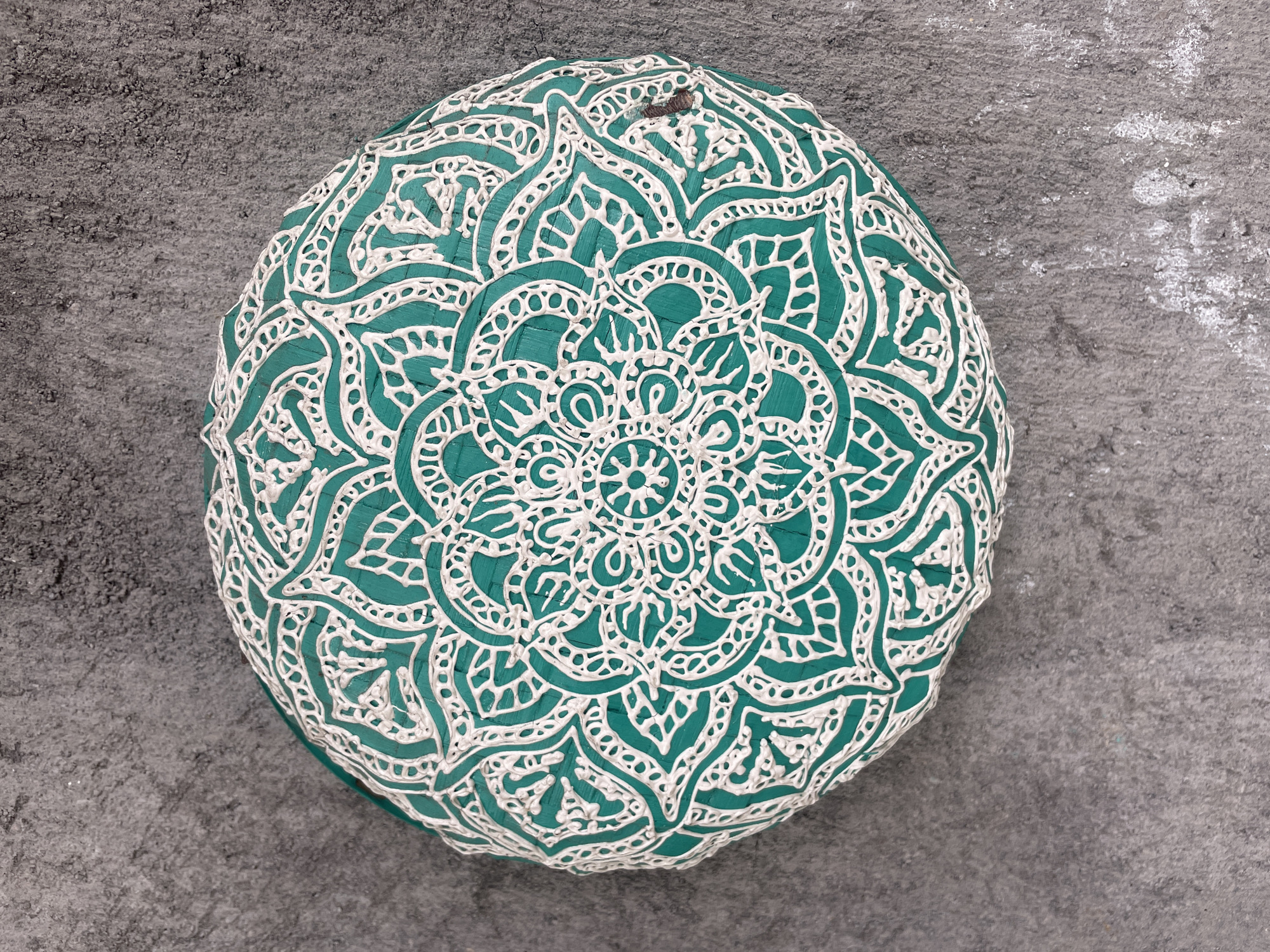 A teal-colored, round cushion with a detailed white mandala design sits on a textured gray surface. The cushion appears slightly three-dimensional, indicating it has some height or volume. The mandala design is intricate, with repeating geometric and floral-like patterns spreading out from the center. The background is a rough, mottled gray, possibly concrete or stone, with some subtle variations in tone and texture. No people are visible in the image. The location is Ubud District, Indonesia.
