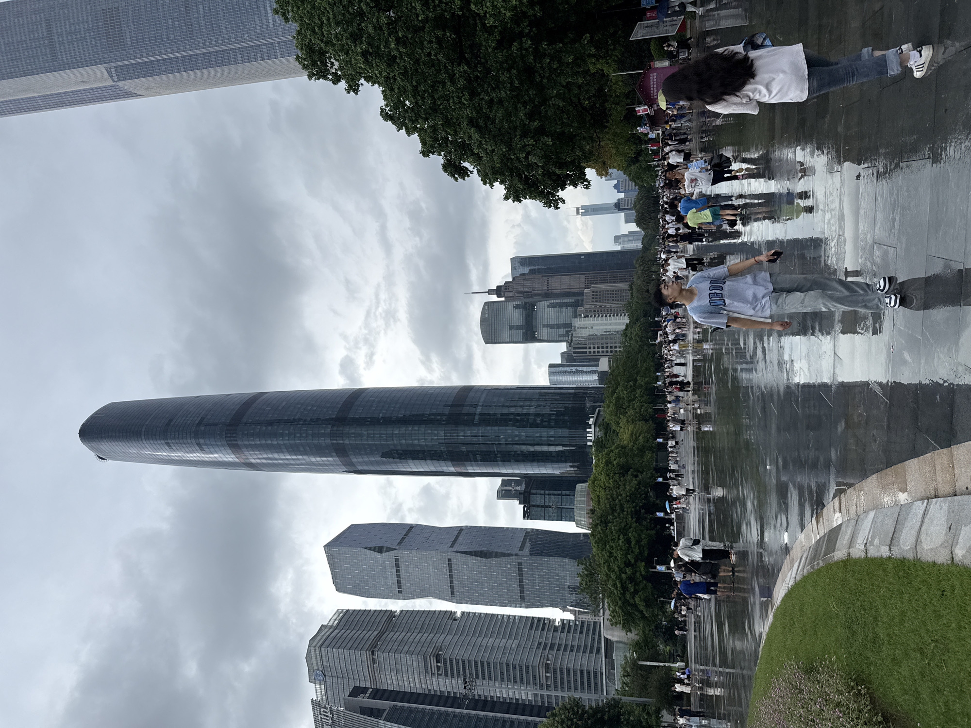 The image depicts a wide, wet, paved plaza in Tianhe District, China, with numerous people walking around. The ground is highly reflective, indicating recent rain. Tall, modern skyscrapers dominate the background, including a distinctive, curved, dark glass tower and other angular, glass-facaded buildings. Green trees are visible along the right side of the plaza and in the mid-ground, interspersed with more trees and greenery in the far distance. The sky is overcast and cloudy. In the foreground, a young man in a light blue t-shirt and grey pants is walking towards the viewer, holding a phone. To his right and slightly ahead, a young woman with long dark hair, wearing a white t-shirt and blue jeans, is walking away from the viewer. Other individuals and groups of people are scattered throughout the plaza, some with umbrellas. The scene suggests a public gathering or a popular urban space.