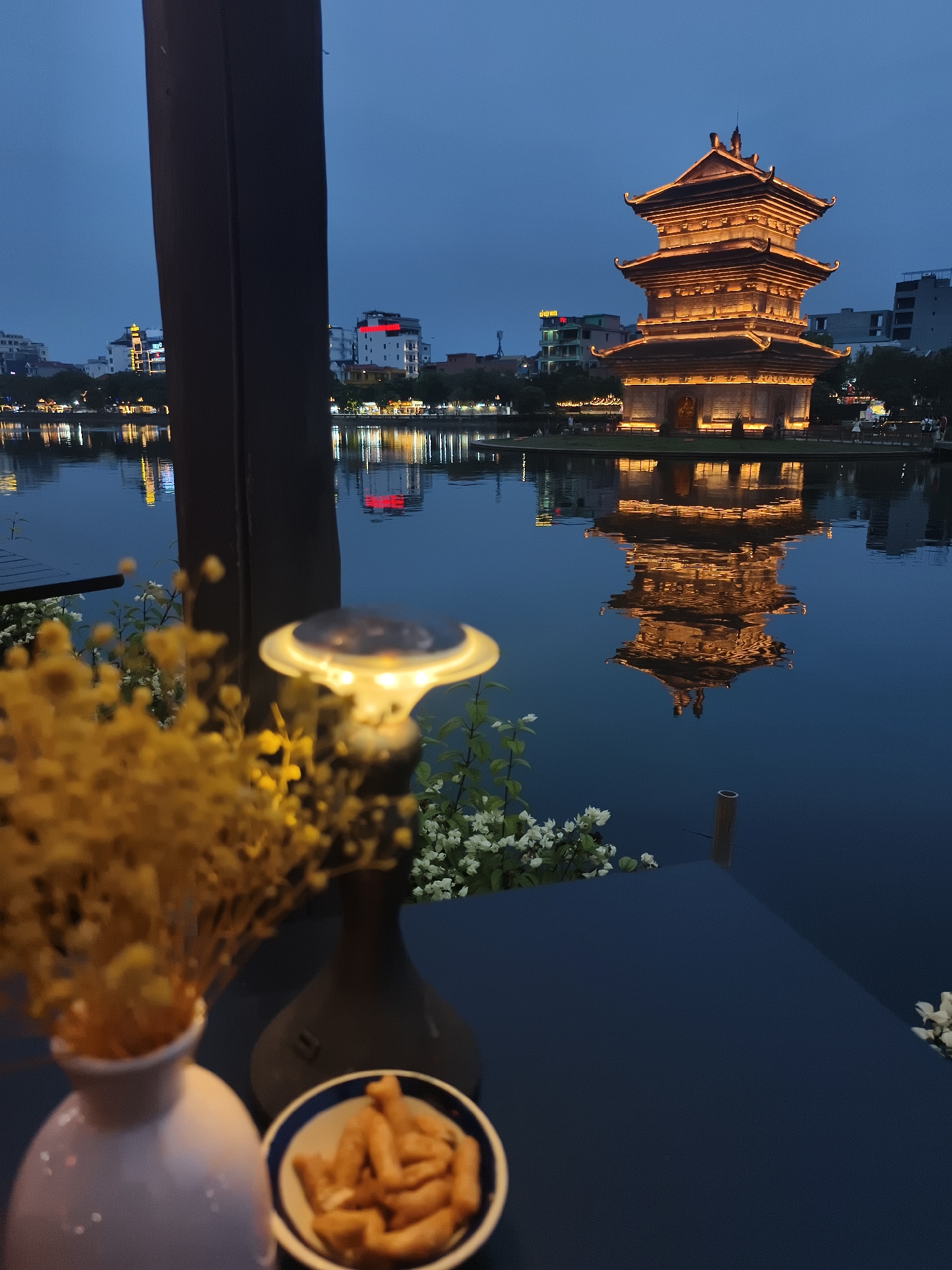 This image captures an evening scene at the Turtle Tower (Thap Rua) in Hoan Kiem Lake, Hoa Lư Ward, Vietnam. In the foreground, a dark, textured surface suggests a table or railing, partially obscured by a vase of dried yellow flowers and a small bowl of snacks. A solar-powered lamp with a bright light is also visible. Beyond this, the calm surface of Hoan Kiem Lake reflects the illuminated Turtle Tower, a multi-tiered structure with ornate architecture, aglow with warm light against the deep blue twilight sky. The distant city skyline features buildings with various lights.