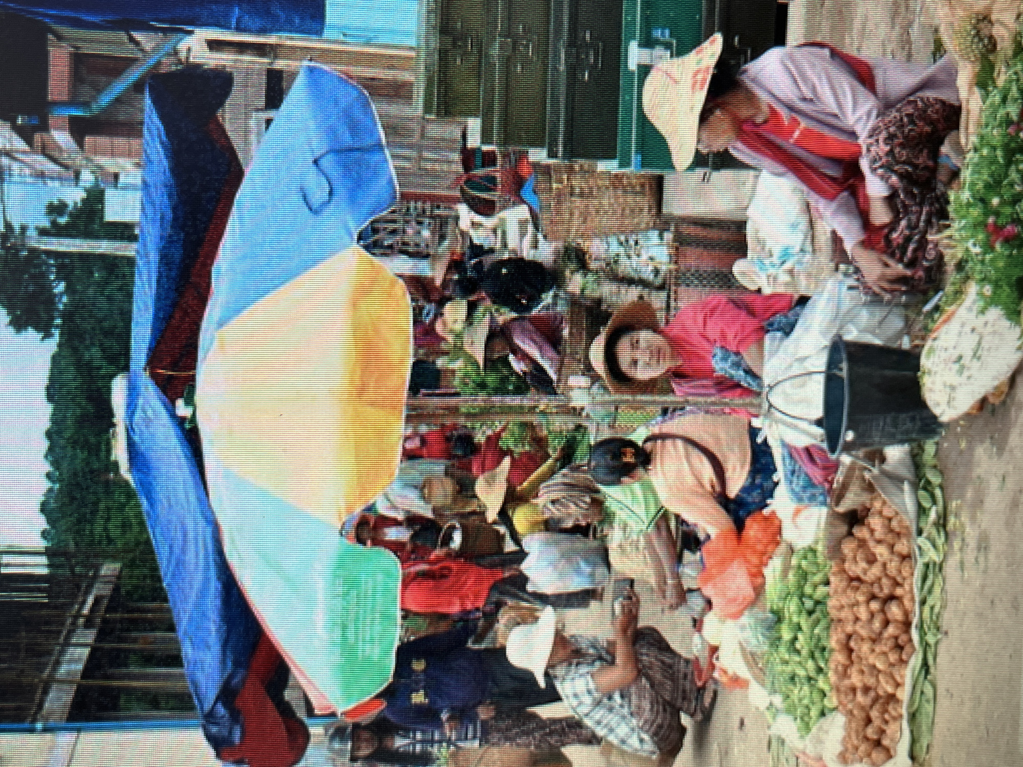 This is a forensic, matter-of-fact description of a street market scene. The location is identified as the city of Sejahtera, Indonesia.
**People:**
Several individuals are present, predominantly women, engaged in market activities. They are dressed in traditional clothing, including brightly colored shirts and patterned skirts or pants. Some wear hats, likely for sun protection. Their postures suggest engagement in selling, buying, or arranging goods. One woman in the foreground is seated with her hands on a pile of produce, while another is crouched and appears to be interacting with items on the ground.
**Objects:**
The scene is filled with a variety of objects typical of a market. Prominently displayed are piles of fresh produce, including what appear to be vegetables and fruits. Bags, some plastic and others woven, are scattered throughout the scene. Large, colorful umbrellas provide shade. Stacked green metal boxes are visible on the right side of the frame, suggesting storage or transport. A black bucket is also present.
**Environment:**
The market appears to be an outdoor setting, possibly a street or an open-air plaza. The ground is paved or dirt. In the background, there are structures that suggest buildings or stalls, and vegetation, possibly trees. The lighting indicates it is daytime.
**Actions and Interactions:**
The individuals are engaged in typical market transactions and preparations. This includes arranging produce, potentially selling items, and interacting with customers or fellow vendors. The overall atmosphere is one of active commerce.