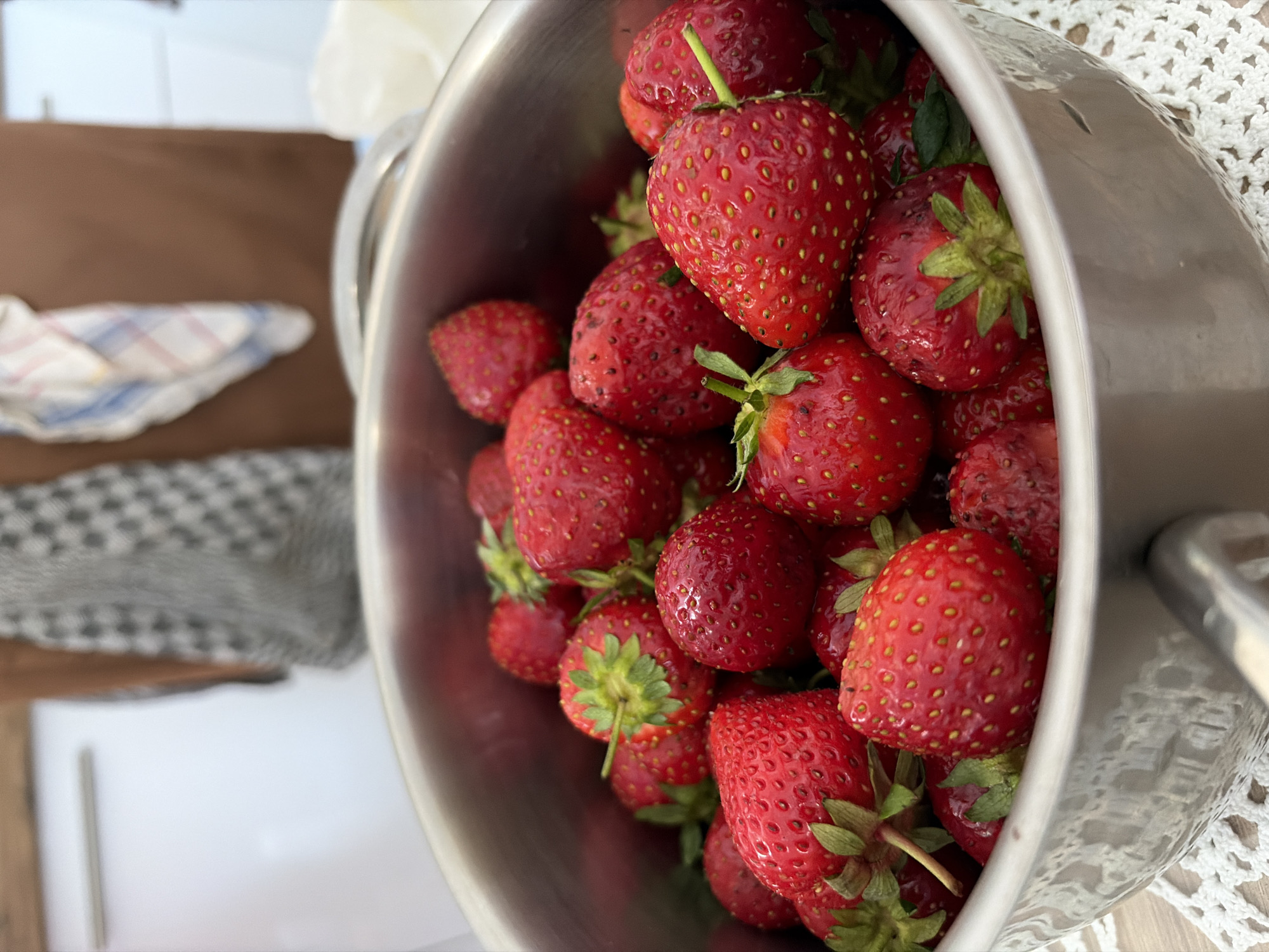 The scene depicts a close-up view of a metal cooking pot brimming with fresh, ripe red strawberries, positioned on a white crocheted lace doily on a light wooden surface in the city of Budva, Montenegro. The strawberries are mostly intact, many displaying their green calyxes, and appear vibrant and moist. The pot is made of a reflective metallic material, possibly stainless steel. The background is significantly blurred, showing elements characteristic of a home kitchen. A person wearing a brown apron is partially visible in the blurred mid-ground. Hanging near the person are two distinct fabrics: a black and white checkered pattern to the viewer's left of the person, and a white, blue, and red plaid towel to the viewer's right. Blurred white kitchen cabinetry or appliances are visible on both the left and right sides of the background. No explicit actions or interactions are depicted, but the arrangement suggests preparation or immediate use of the strawberries.