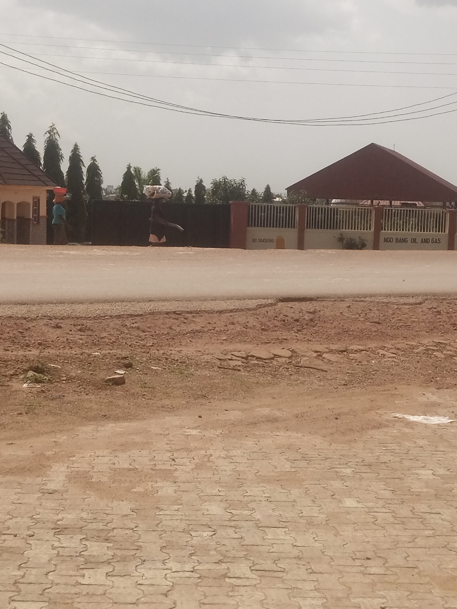 The image shows an outdoor scene with two people and a building in the background. A person on the left is wearing a blue shirt and striped pants, carrying something on their head. Another person on the right, wearing a dark top and a skirt, is also carrying a load on their head. They appear to be walking along a dirt road in front of a dark fence. Behind the fence is a building with a red roof and a white fence. There are several tall, dark green trees in the background, and some power lines are visible in the sky. The ground in the foreground is made of interlocked pavers. The weather appears overcast, suggesting it might be daytime. Visible text on the building reads "NO SMOKING" and "NGO BANG OIL AND GAS". The overall scene suggests a commercial or industrial area with people engaged in manual labor.