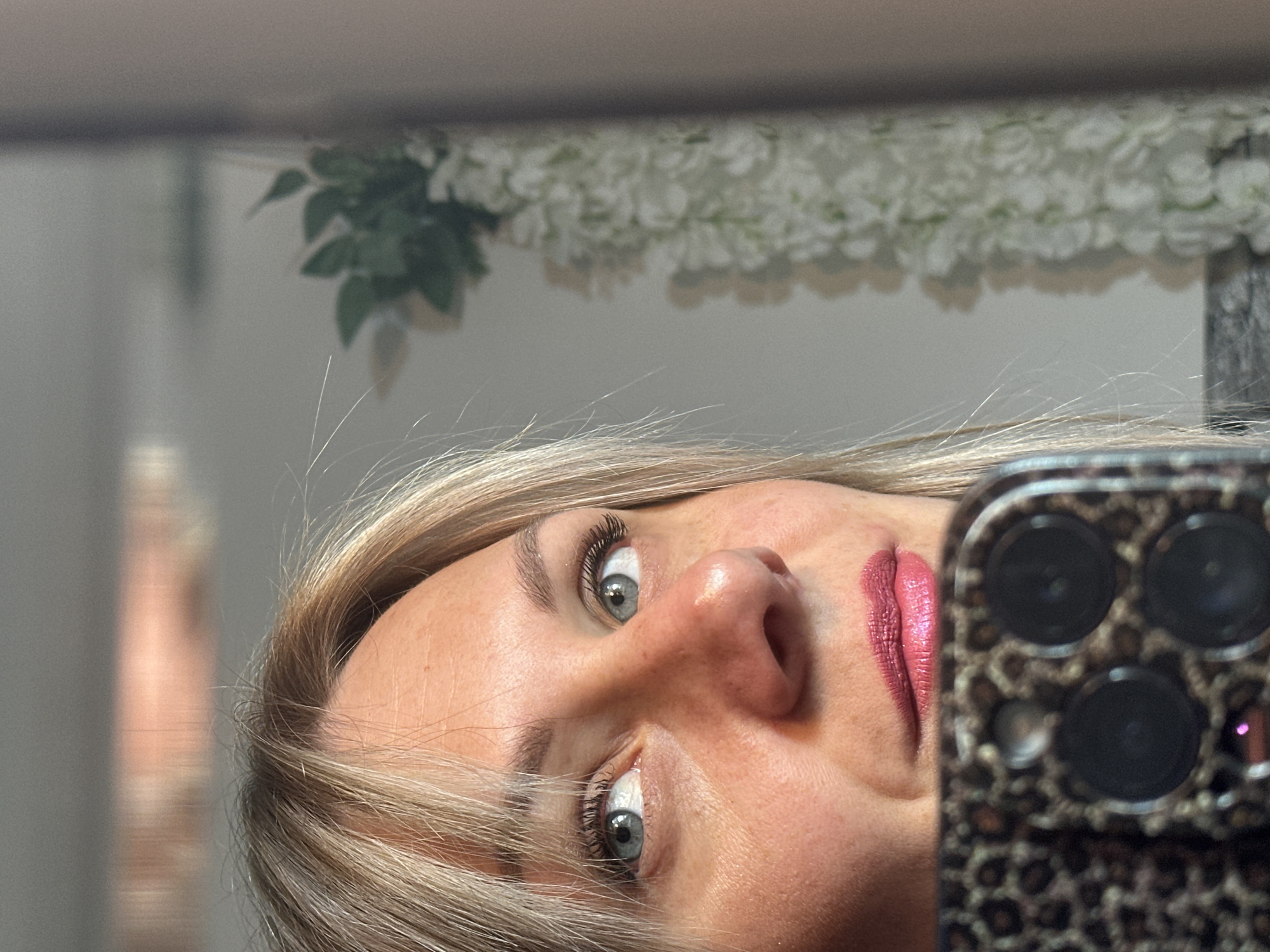 A blonde woman with blue eyes is taking a selfie in a mirror. She is holding a phone with a leopard print case. Her lips are a reddish-pink color. In the reflection of the mirror, white flowers are visible along with a partial view of a doorway. There are no specific details indicating that this image was taken in Sesto Campano, Italy.