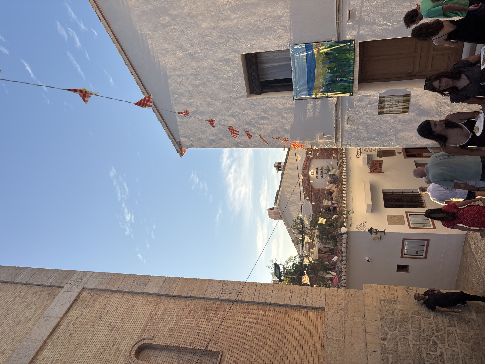 This image depicts an outdoor gathering in a narrow street, likely in a Spanish town given the architectural style and context. The time of day appears to be late afternoon or early evening, with warm, directional sunlight casting long shadows and highlighting the whitewashed buildings. The sky is a clear blue with a few wispy clouds.
Several people are visible, dressed casually, suggesting a social event. Some are standing and conversing, while others appear to be seated or mingling. There are strings of triangular flags or pennants strung across the street, adding a festive atmosphere. One building has a colorful mural or banner hanging on its exterior. Signs are visible on some of the buildings, including one that reads "EL CANONGE TERRAZA" and another that seems to be "LE GOUACHE RESTAURANT". There's also a sign that says "TRENDY TREE".
The scene suggests a relaxed community event, possibly a street festival or a casual outdoor dining experience. The overall mood seems convivial and pleasant, with people enjoying the warm weather and each other's company. The architecture, with its whitewashed walls and terracotta roof tiles, is characteristic of Mediterranean towns.