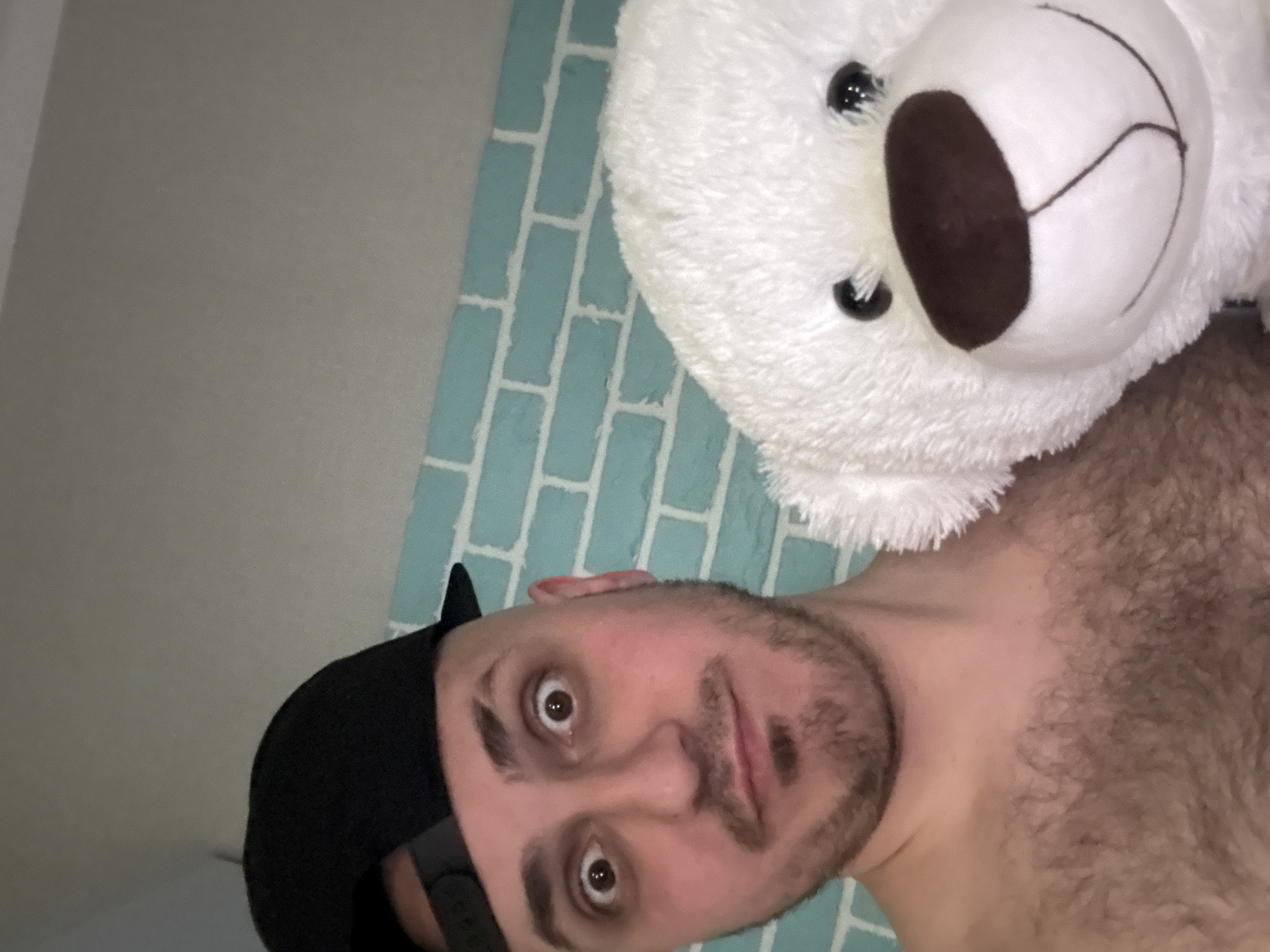 A man with a black baseball cap is lying on a light blue, brick-patterned surface next to a large, white teddy bear. The man has brown eyes and a short beard. The location is indeterminate and cannot be inferred from the image.