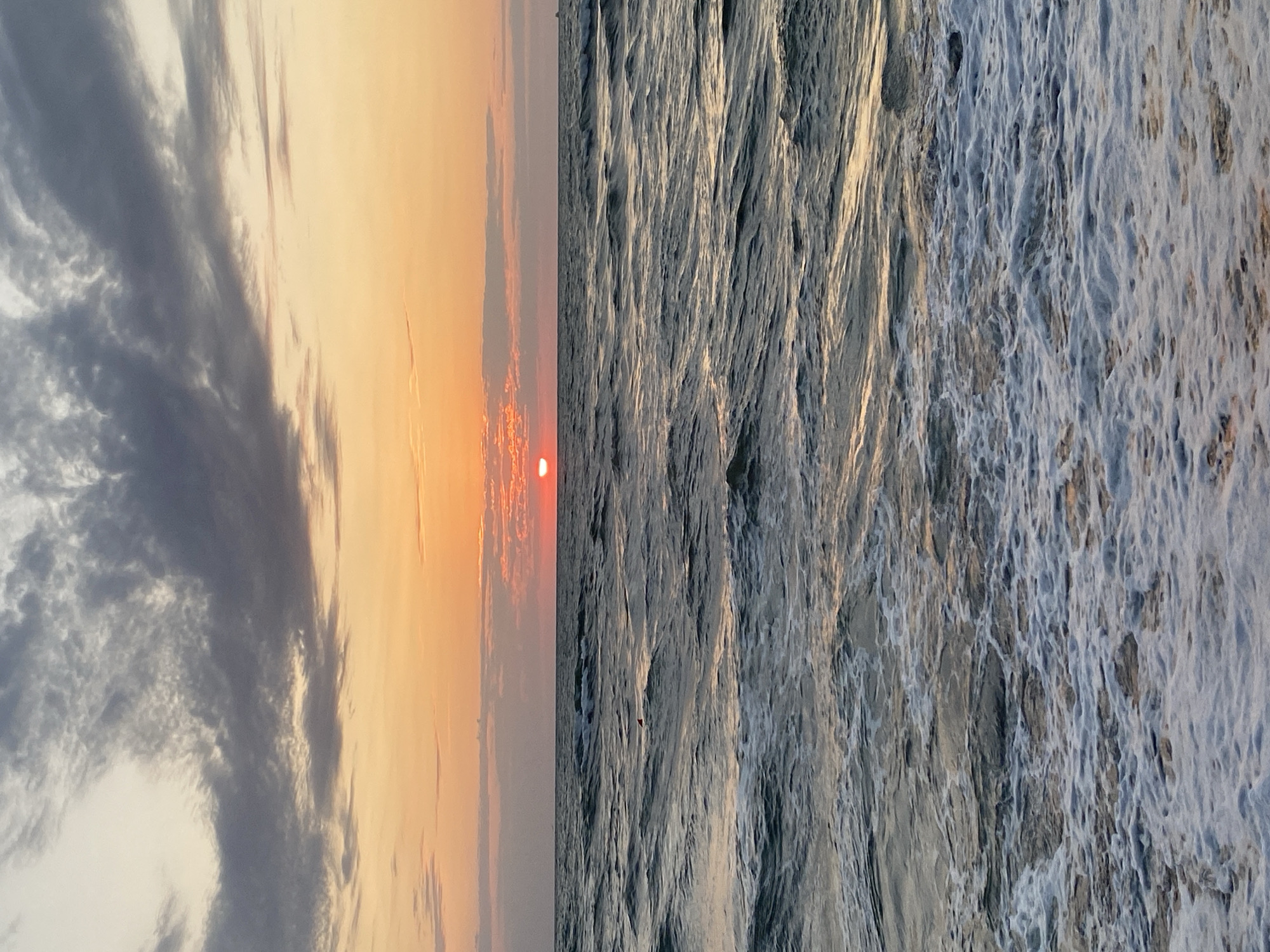 A vertical, low-angle shot captures a sunset over the Black Sea in Ureki, Georgia. The sky is a gradient of soft orange and peach hues, transitioning into muted gray clouds with pink undertones. The sun, a vibrant red orb, is partially obscured by a thin band of clouds near the horizon. The sea is a deep, textured blue-gray, with white foam and waves crashing at the shore in the foreground. A small red buoy with a triangular top is visible in the middle ground, floating on the water. In the far distance, a faint silhouette of a ship can be seen on the horizon. The overall mood is tranquil and serene, typical of a seaside sunset.
