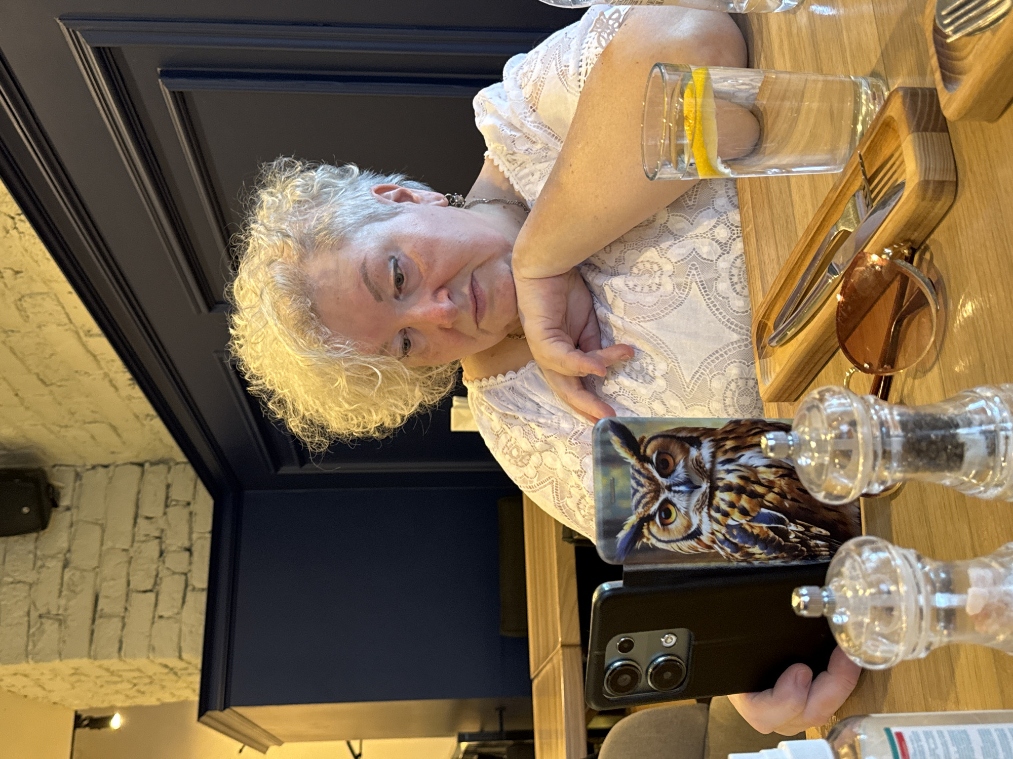A woman with short, curly, blonde hair is sitting at a wooden table, looking at her smartphone. She is wearing a white lace top and a necklace. Her left elbow is resting on the table, and her head is propped on her hand. In front of her are two salt and pepper grinders, a pair of sunglasses, a wooden utensil holder with a knife and fork, and a glass of water with lemon slices. The phone has a case with a picture of an owl on it. In the background, there is a dark blue paneled wall with white brick above it. The scene appears to be in a restaurant, possibly in Kazan, Russia, given the context.