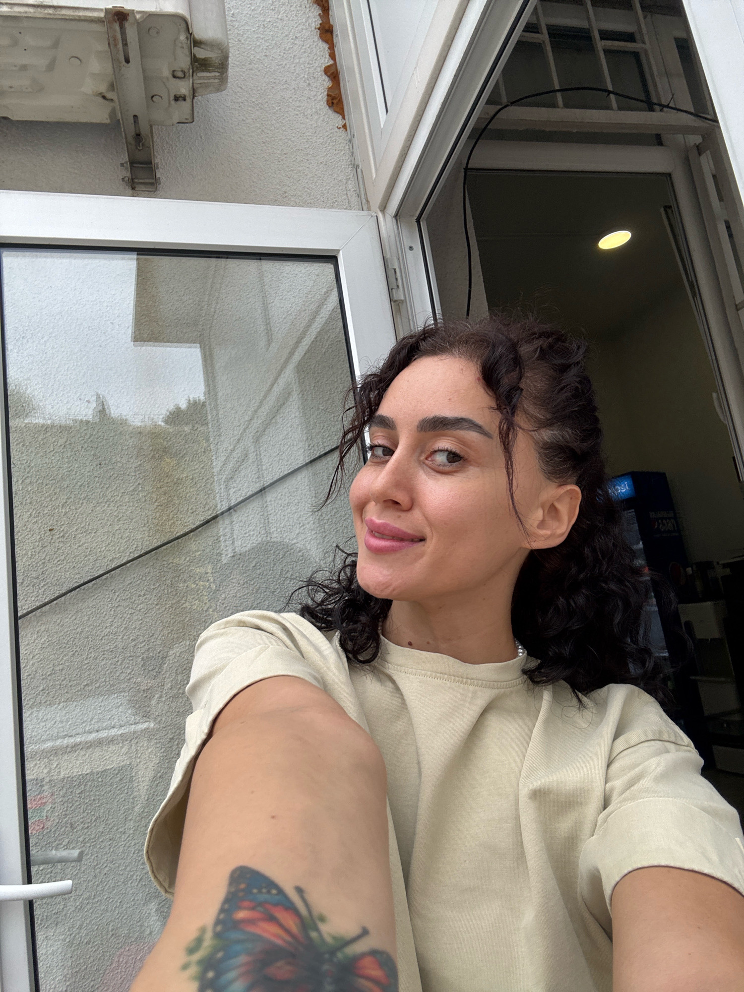 A woman with curly dark hair and fair skin is taking a selfie in what appears to be an outdoor setting, possibly on a balcony or near a window. She is wearing a light beige t-shirt and has a butterfly tattoo on her arm. In the background, there is a white wall with some texture, a window with a white frame, and an open door revealing a dark interior with a bright light fixture and a blue Pepsi-branded refrigerator. The scene is likely in Tbilisi, Georgia, given the context. The lighting suggests it might be overcast or late afternoon.