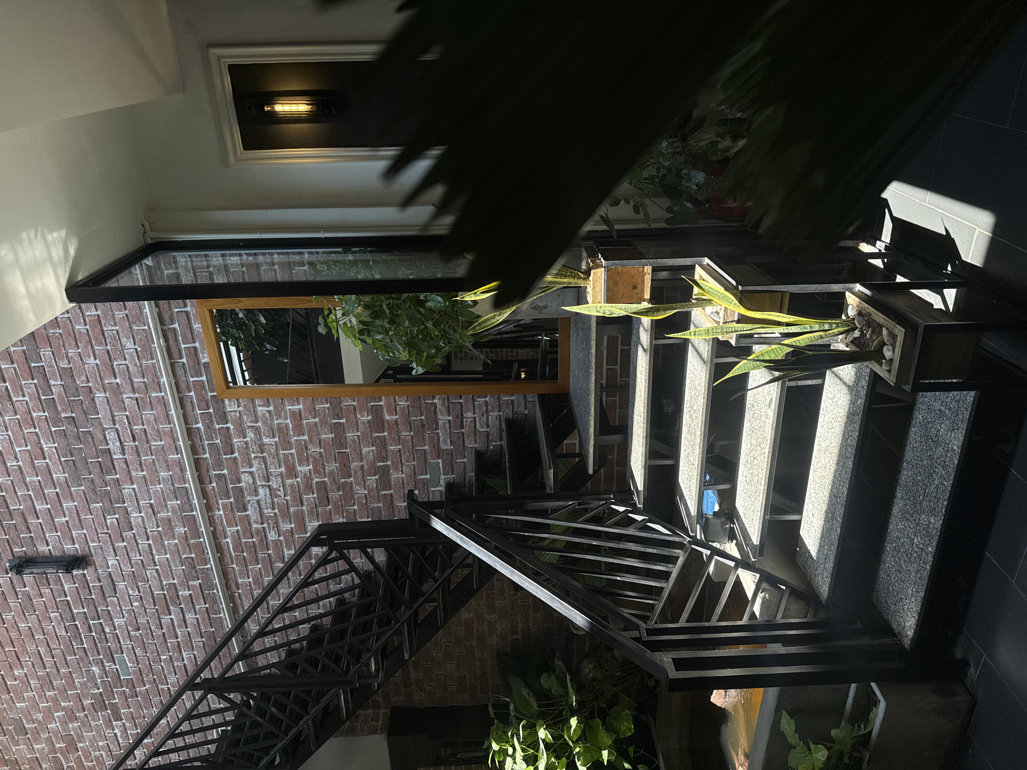 The image shows an interior space featuring a staircase with black metal railings. The staircase has grey steps, with some sections appearing to be concrete or stone. The left wall is made of red brick and includes a rectangular window. There are plants located near the staircase, some on the steps themselves. Lighting fixtures are visible on the walls and ceiling. The location of the image is Unknown, Unknown. There are no people visible in the image.
