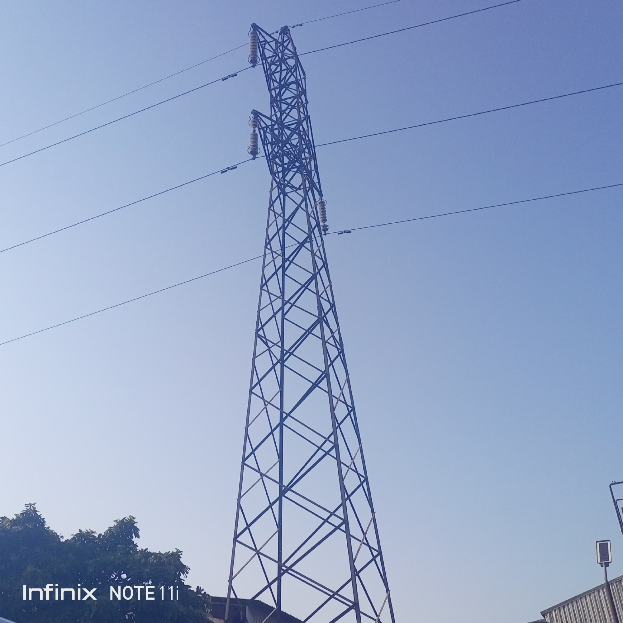 A power transmission tower stands against a clear blue sky. The tower is constructed of a dark metal lattice and supports three levels of horizontal power lines. At the base of the tower, foliage and a structure are visible. The location is Idemili, Nigeria. A logo for "Infinix NOTE 11i" appears in the lower-left corner of the image.