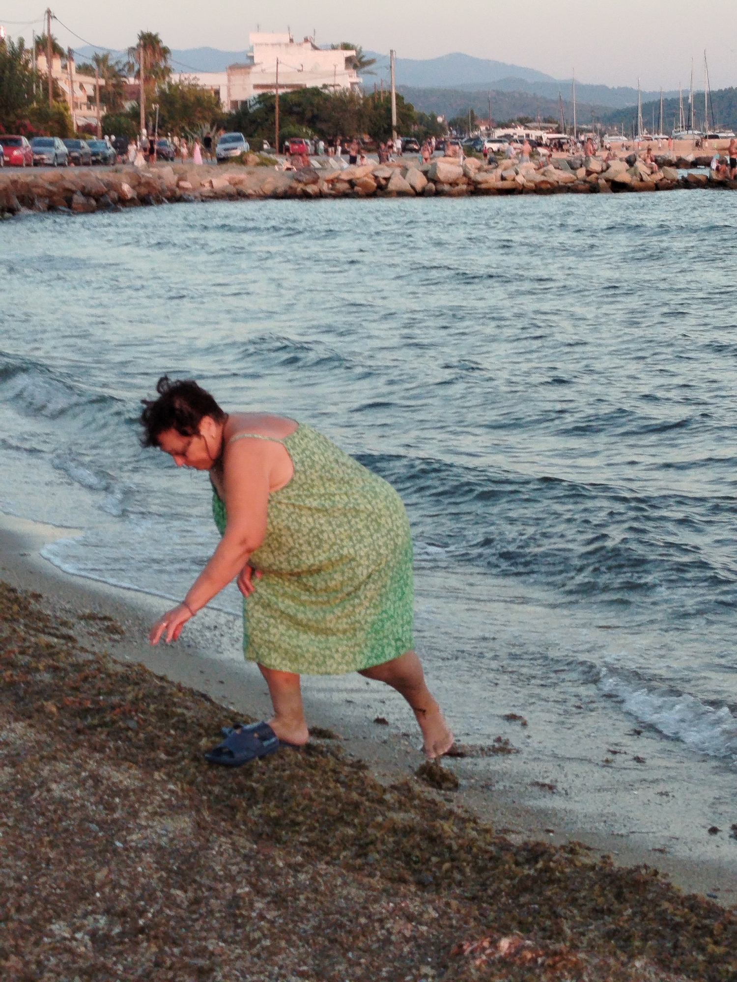 A woman in a green floral print dress is bent over on a beach in Sithonia Municipal Unit, Greece, reaching her hand towards the wet sand and seaweed at the water's edge. Her right foot is bare and rests on the shore, while a dark sandal lies nearby on the sand. The sea is rippling with moderate waves. In the background, a shoreline with rocks and a walkway is visible, with parked cars, buildings, palm trees, and a harbor with numerous sailboats. The sky is overcast.