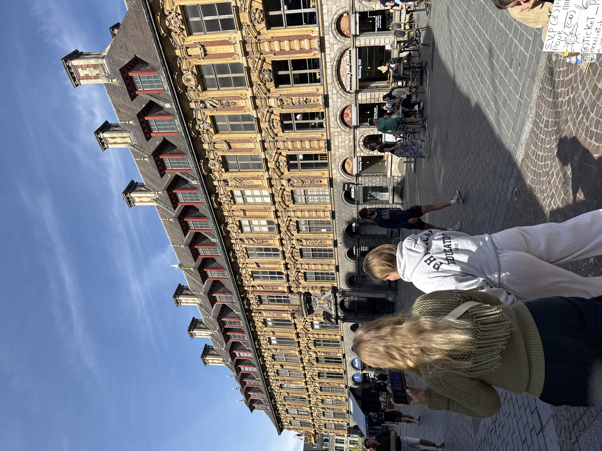 The image depicts a sunny daytime scene in the Grand Place of Lille, France. The architecture of the surrounding buildings is ornate and distinctly Flemish Baroque style, with a large, elaborate building dominating the background. This building features a yellow facade adorned with intricate carvings, numerous windows, and a steep, tiled roof with dormer windows and chimneys. In the foreground, two women are walking away from the camera. The woman on the left has blonde hair and is wearing a green knitted sweater and dark pants, holding a smartphone. The woman on the right has lighter blonde hair and is wearing a grey hooded sweatshirt and light-colored pants. Other people are visible in the middle ground, some sitting at outdoor cafe tables. The ground is paved with cobblestones, and the bright sunlight casts sharp shadows. Signs for businesses such as "Notting Hill Coffee" are visible.