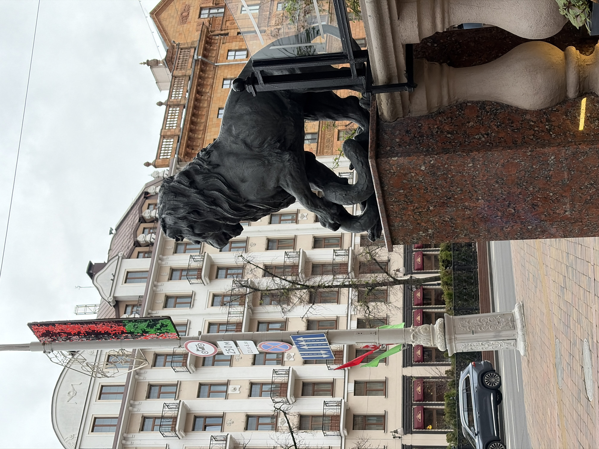 The image captures a street scene in an urban environment. A bronze lion statue stands prominently atop a red granite pedestal. Behind the statue, a tall building with multiple windows and balconies can be seen. Street signs are visible on a pole, and a black SUV is parked along the brick-paved sidewalk. The sky is overcast.