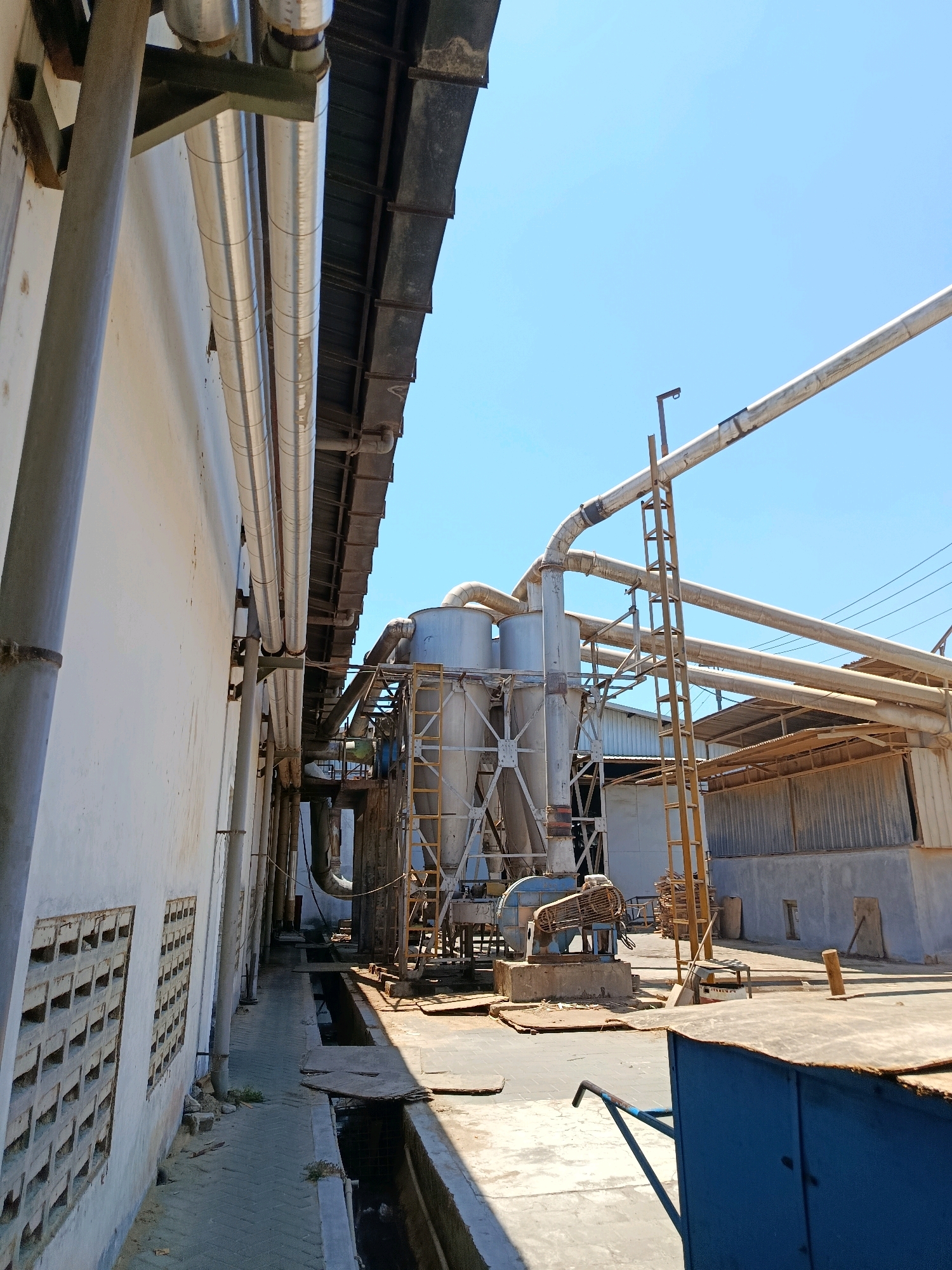 The image depicts an industrial area in Kembangarum, Indonesia, likely a factory or processing plant. The scene is dominated by large, metal pipes running along a white building on the left and extending overhead. These pipes are insulated and appear to be part of a ventilation or material transport system.
In the background, a complex industrial structure with multiple cylindrical tanks is visible, supported by a metal framework. A yellow ladder leads up to this structure, suggesting access for maintenance or operation. Next to the tanks, a large fan or blower unit is mounted on a concrete base.
The ground surface is a mixture of concrete and paved areas, with a visible drainage channel running alongside the building. Debris and some vegetation are present. The environment is outdoors, under a clear blue sky, indicating daytime. The overall impression is one of an active industrial site. No people are visible in the image.