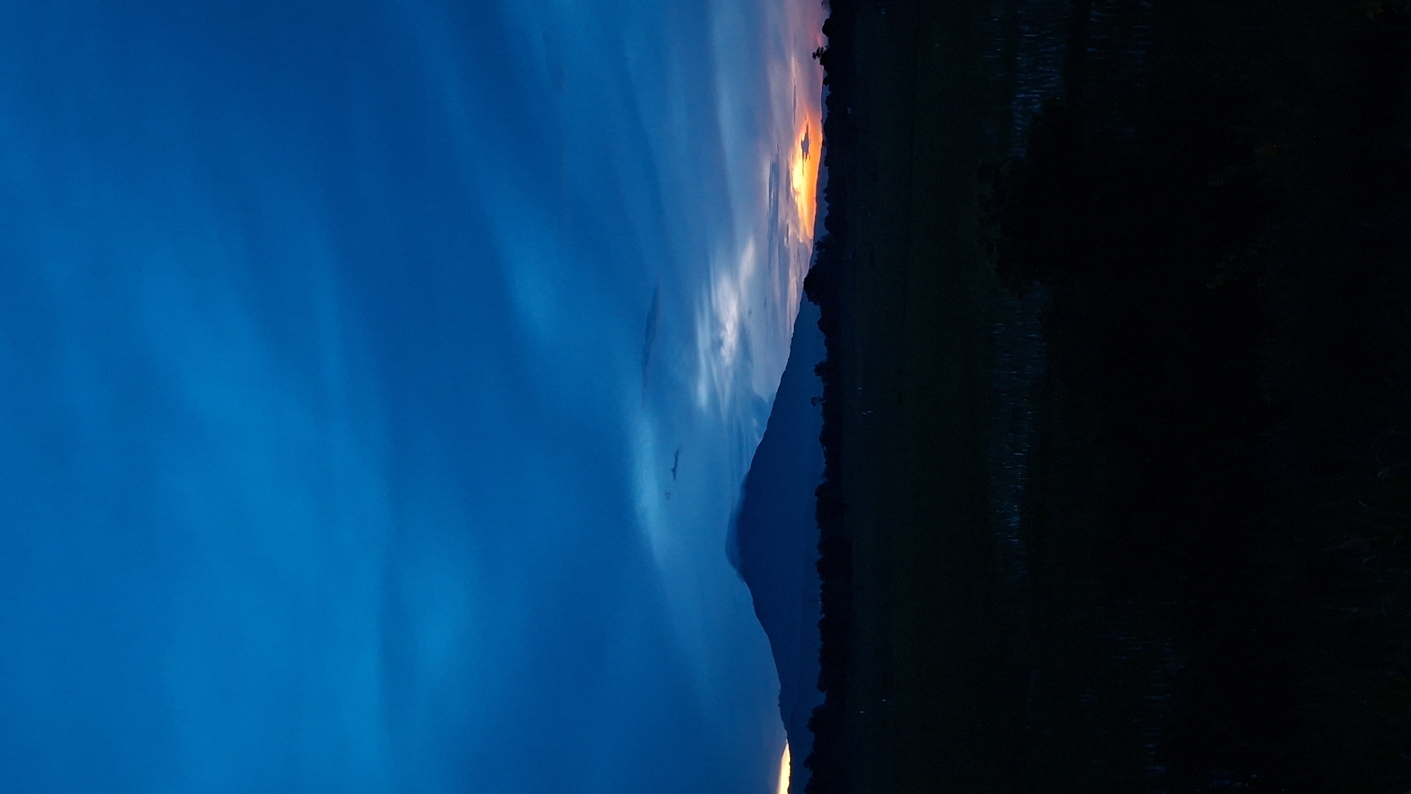A solitary, dark mountain peak rises from a silhouette of trees in the middle ground of the image, shrouded in a light cloud near its summit. The sky above is a deep, rich blue, with streaks of lighter blue and grey clouds. To the right of the mountain, a sliver of orange and yellow light pierces through the horizon, suggesting a sunset or sunrise. The foreground is a dark, indistinct field, possibly a rice paddy, with some vegetation visible. The overall scene is serene and atmospheric, indicative of dusk or dawn in a rural setting. The image appears to depict the natural landscape near Baksukon, Indonesia, where such mountainous terrain and agricultural fields are common.