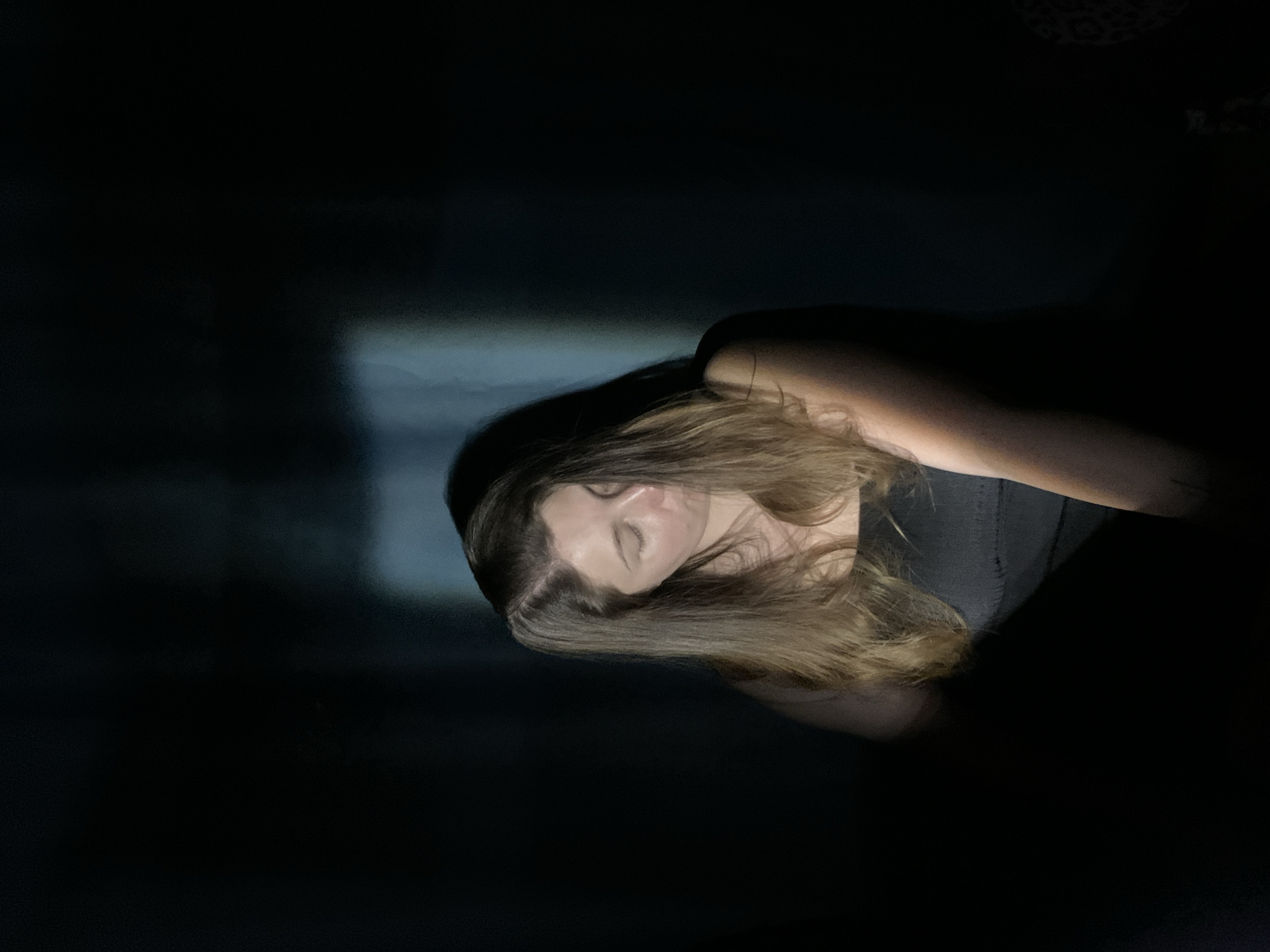 The image presents a person with long brown hair lying down, seemingly in a dimly lit room. A faint light source illuminates part of their face and hair. The person appears to be wearing a dark-colored top. The background is largely obscured by darkness, with a vague, blurry shape possibly indicating a window or wall. There is nothing in the image to suggest that it was taken in Chaikovsky, Russia. The subject is lying down.