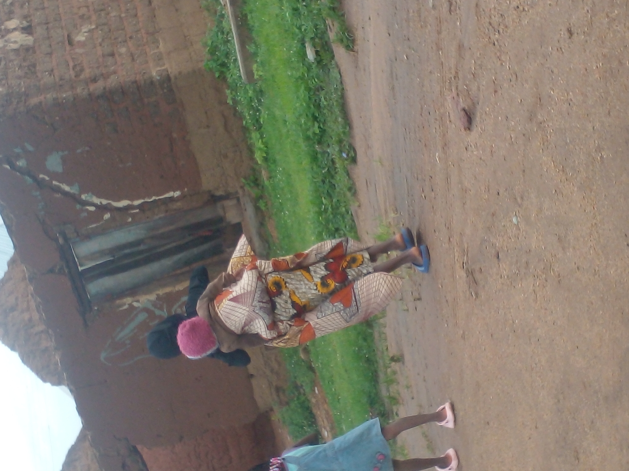 In Langtang, Nigeria, two young girls are the subjects of this outdoor photograph. The girl on the left wears a light blue dress. The girl on the right carries another child on her back, who is wearing a pink hat and a patterned wrap. The background shows a rustic, mud-brick structure, likely a home or building, with a corrugated metal door. Green grass grows in the background, and the ground in the foreground is dirt. The image captures a moment of everyday life in this Nigerian community.