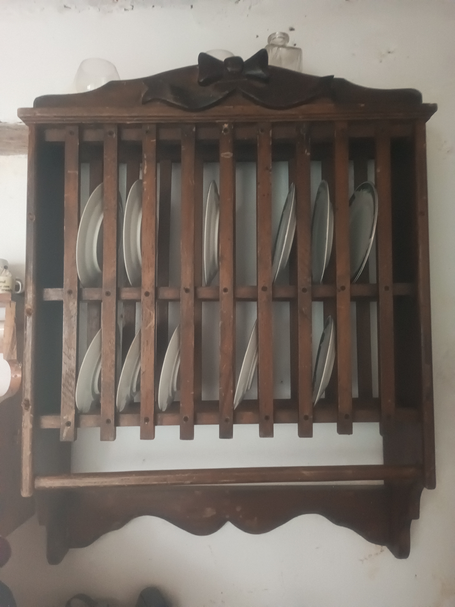 A vintage wooden plate rack hangs on a white wall in what appears to be a home or establishment in Juan Griego, Venezuela. The rack is filled with approximately 15 white plates of various sizes. The top of the rack features decorative wooden carvings. A horizontal wooden bar is situated below the plate slots. The wall above the rack shows signs of age and wear, with some discoloration and chipping. The bottom of the image shows the lower portion of the rack and the wall below it, with a suggestion of a shelf or cabinet underneath the plate rack. No people are visible in the image.