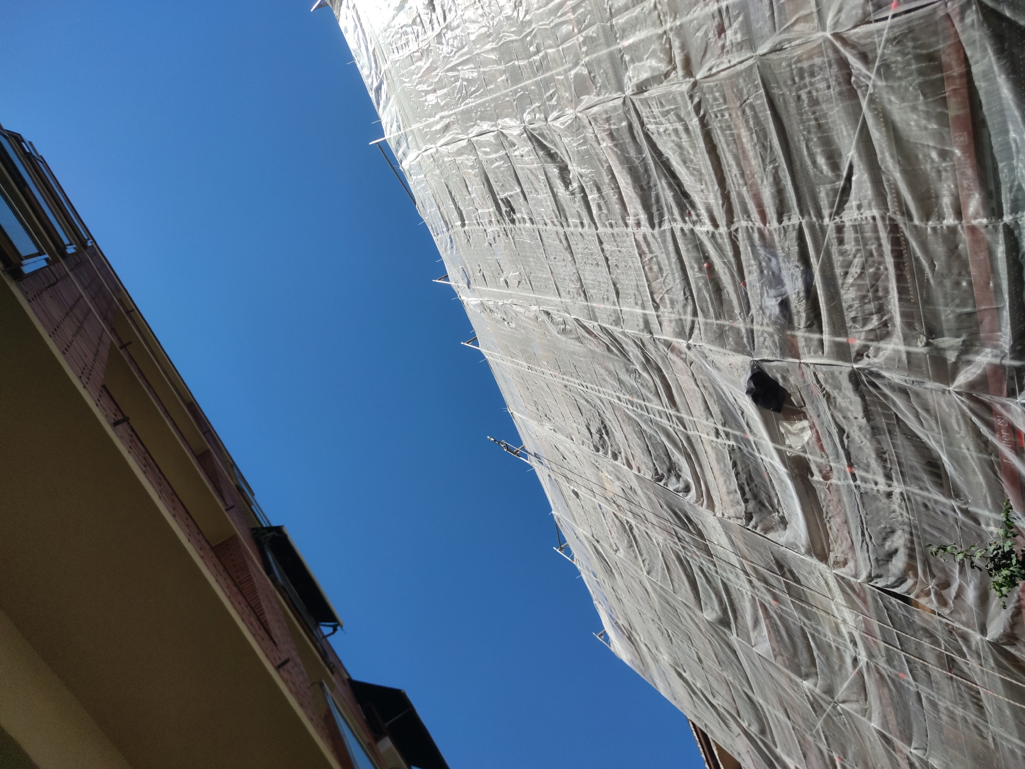 The image shows a low-angle view of two buildings against a clear blue sky. The building on the left is constructed of brick and has a beige exterior. It includes visible windows. The building on the right is shrouded in a white plastic sheeting, likely for construction or renovation. Scaffolding is visible underneath the sheeting. There are visible tie-downs securing the plastic sheeting. A bush or tree branch is visible emerging from the sheeting on the side of the building. The scene is outdoors during daylight hours. The location is a city, but specific city or country cannot be identified.