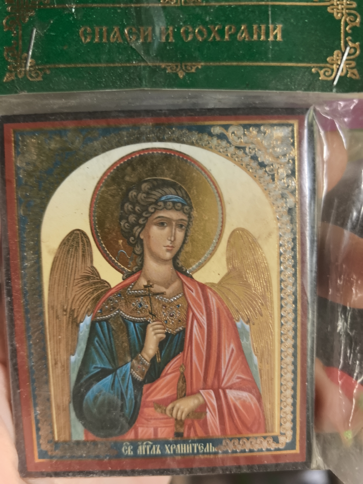The image shows a small, framed Orthodox icon depicting an angel. The angel has dark curly hair with a blue ribbon, a halo, and golden wings. It is wearing a blue robe and a red cloak, and holding a cross in its right hand and a sheathed sword in its left. The icon is bordered with gold ornamentation. The inscription on the top of the packaging reads "Спаси и сохрани" (Save and Protect) and the inscription on the bottom of the icon reads "Святой Ангел Хранитель" (Holy Guardian Angel). The image shows the icon being held in a plastic package. There is no clear indication of a location within the city of Noviy, Russia.
