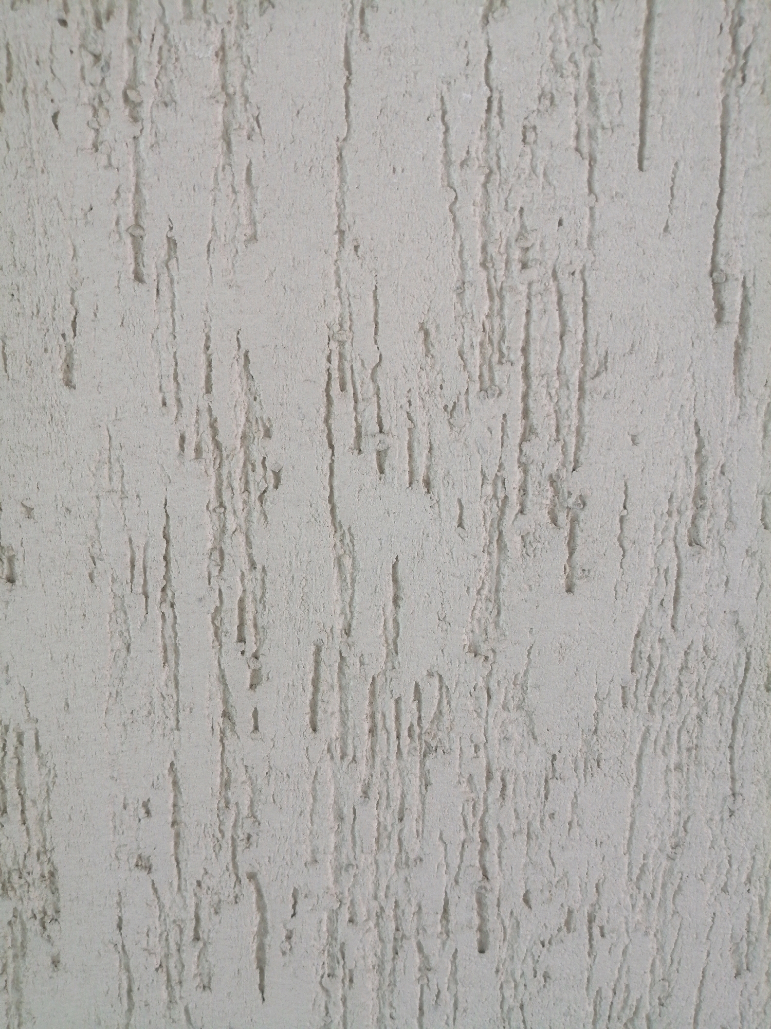 This is a close-up, eye-level, vertical, full-frame, indoor shot of a textured, light-colored wall. The wall has a rough, uneven surface with vertical striations and indentations, creating a pattern that resembles coarse bark or dried mud. The lighting appears to be neutral, with no strong shadows, suggesting it is either overcast or from an artificial source. The color is a pale, off-white or light gray. There are no people, animals, or other objects visible in the image, and the background is entirely composed of this textured wall. The exact location within the city of Unknown, Unknown is not discernible from the image itself.