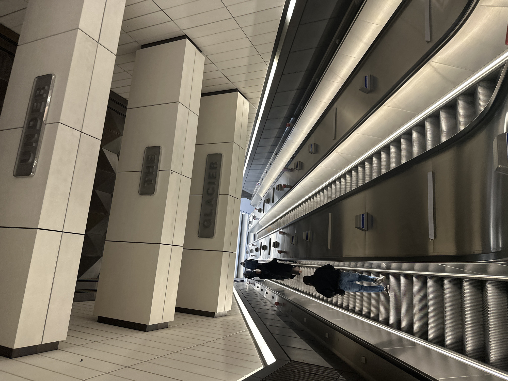 The image depicts a scene in a London Underground station, United Kingdom. The setting features escalators, ascending or descending, flanked by columns with signage indicating station names, possibly including "Under," "The," and "Glacier." People are visible on the escalator, though their features are indistinct. The environment is clean and modern, characterized by white tiled floors and walls. The overall impression is of a well-maintained and functional transport hub.