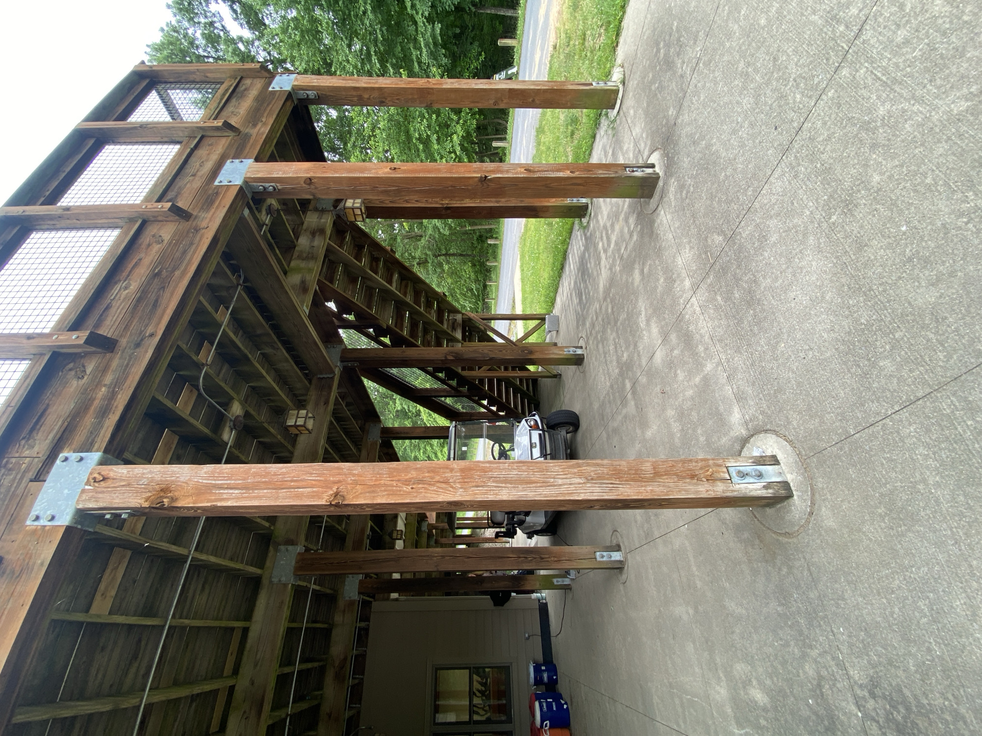 A golf cart is parked under a wooden deck that is supported by large, wooden posts in Washington Township, United States of America. The deck has stairs leading up to it and a railing with wire mesh. To the left of the golf cart, there are several blue and orange buckets. The ground is paved with concrete. In the background, there is a road and a grassy area with trees. The sky is overcast.