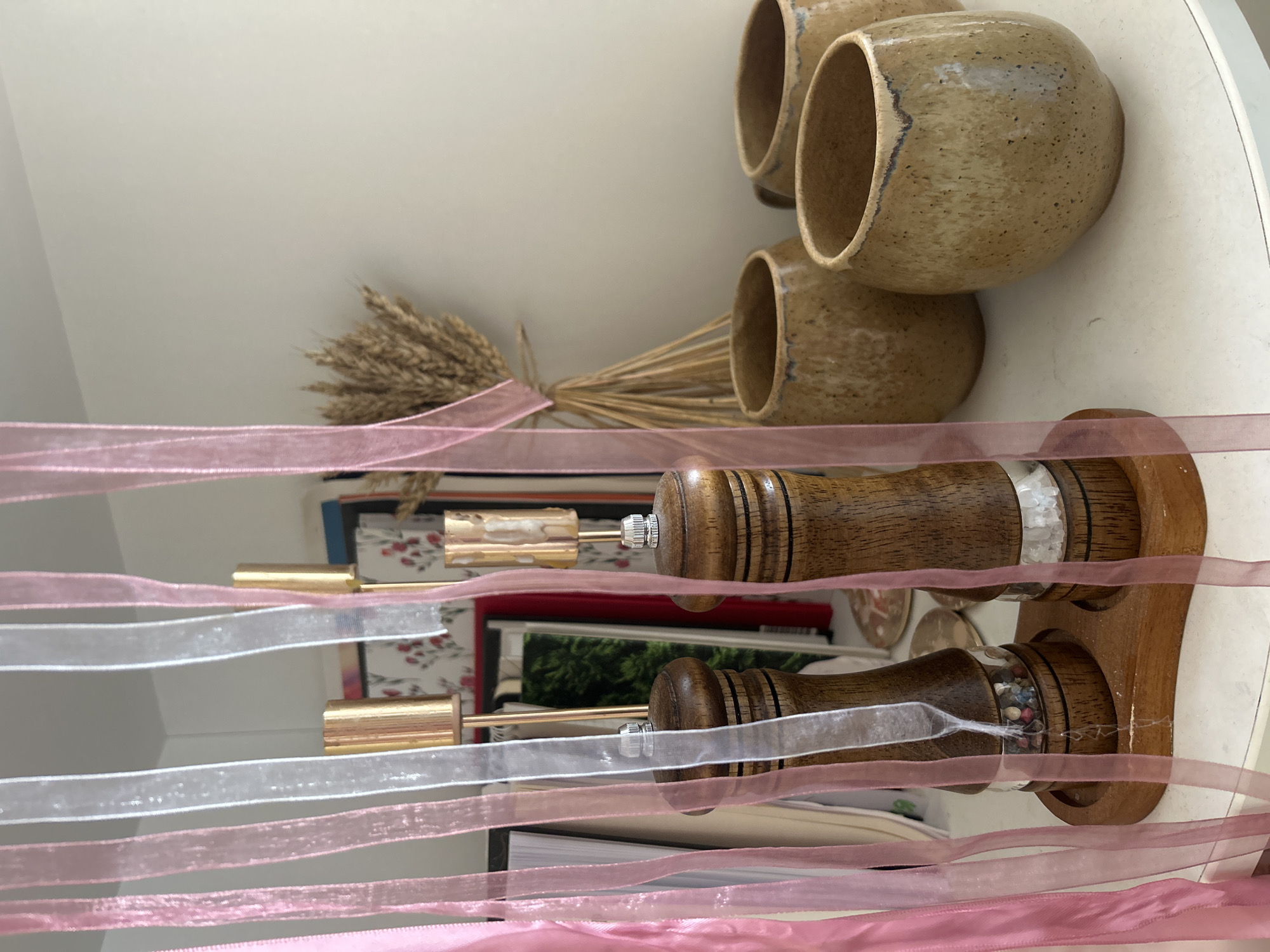 A still life scene is captured on a shelf, featuring several objects. Three ceramic mugs with a grainy texture are positioned on the top right, next to a bouquet of wheat tied with a pink ribbon. Below these items, a stack of books provides a base for a wooden salt and pepper shaker set. Pink and white ribbons are draped across the foreground, partially obscuring the view of the objects behind them. The scene is lit by an unidentified light source from the top. The location is undetermined, with no clear indications of being in Balashikha, Russia.
