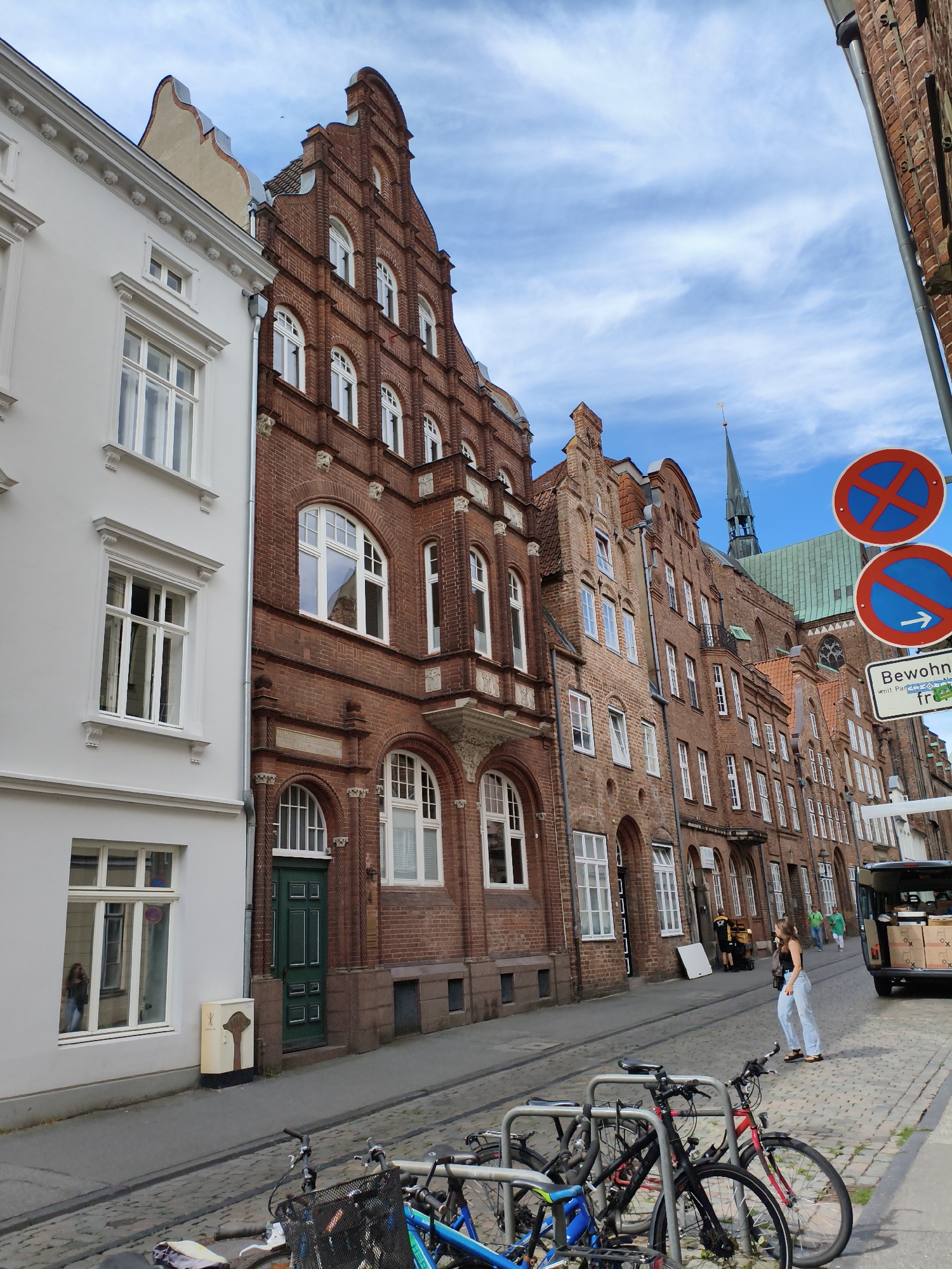 The image depicts a street scene in Lübeck, Germany, characterized by historic brick buildings lining a cobblestone street. On the left, a modern white building with large windows stands in contrast to the older architecture. Several bicycles are parked in the foreground on the right side of the street, some with baskets. Further down the street, a woman in light blue jeans and a dark top is walking towards the right. She is on the sidewalk next to parked bicycles and a white van filled with boxes. Traffic signs, including two "no parking" signs and a directional sign, are visible on the right side of the street. The sky is blue with scattered white clouds. A church spire is visible in the distance, rising above the rooftops of the buildings. The overall impression is of a well-preserved historic urban environment.