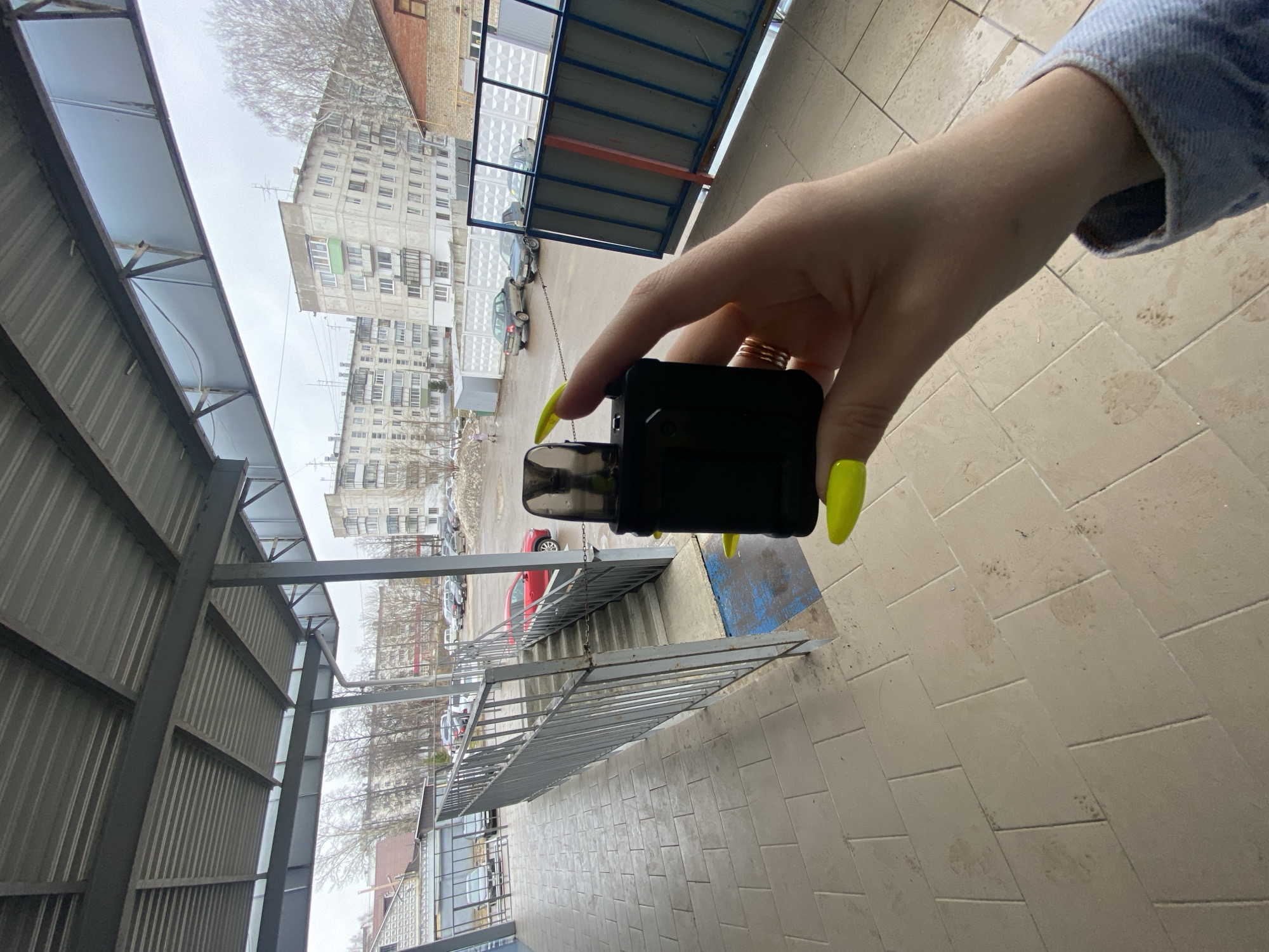 A person is holding a black vaping device outdoors in Chaikovsky, Russia. The person's hand, with bright yellow manicured nails and several rings on the fingers, is visible. The person is wearing a denim jacket. In the background, there are apartment buildings, a covered walkway structure, parked cars, and a metal railing. The ground is covered with light-colored tiles. The sky is overcast.