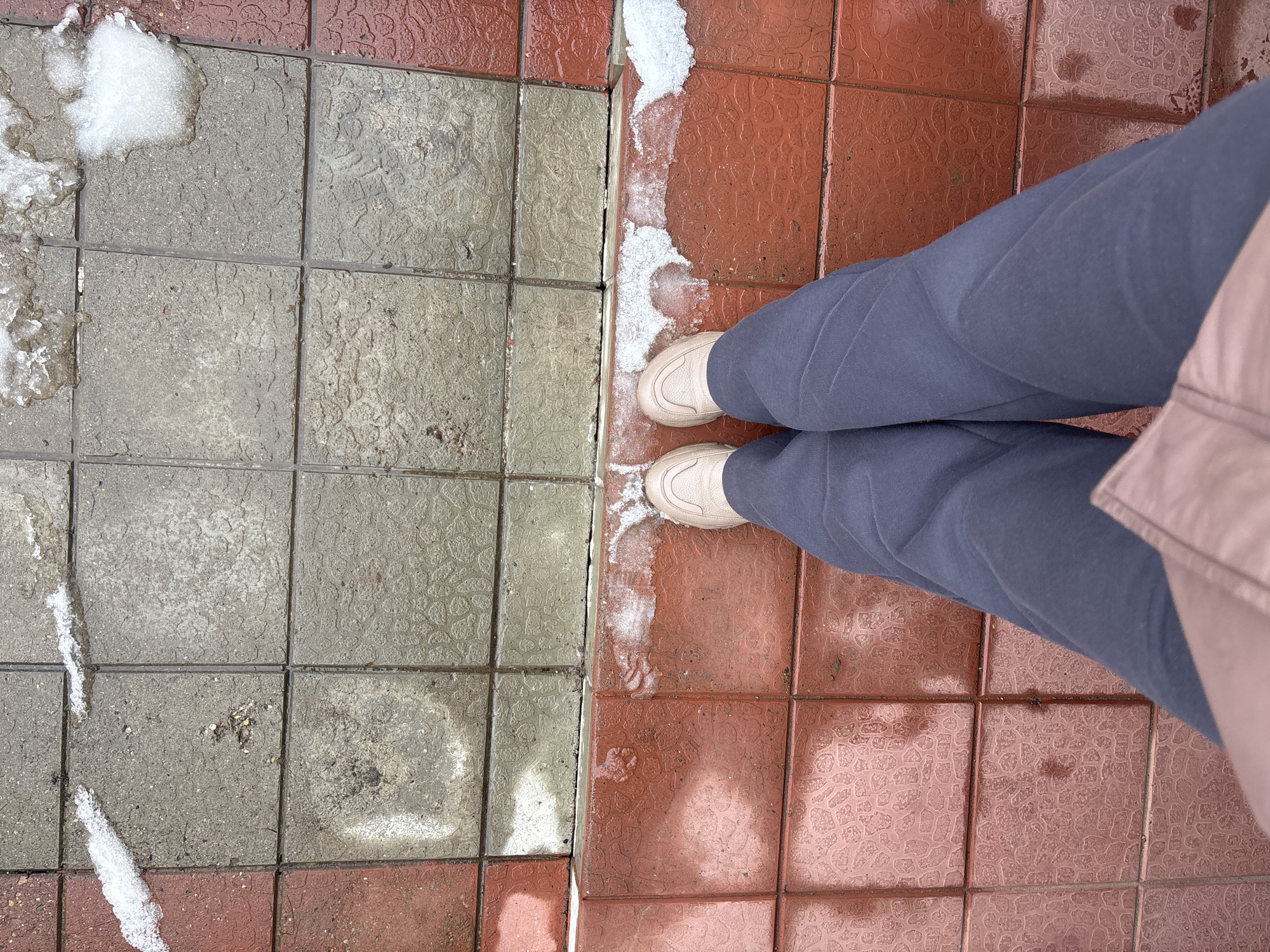 A high-angle shot depicts a person standing on a tiled surface, potentially a sidewalk, in Ферма, Russia. The tiles are divided into two distinct color groups: gray tiles on the left and red tiles on the right. Patches of melting snow are present on both tile colors, primarily along the divide and in the corners. The person is wearing a pair of beige sneakers and blue sweatpants. A portion of a light pink jacket is visible on the right edge of the image.