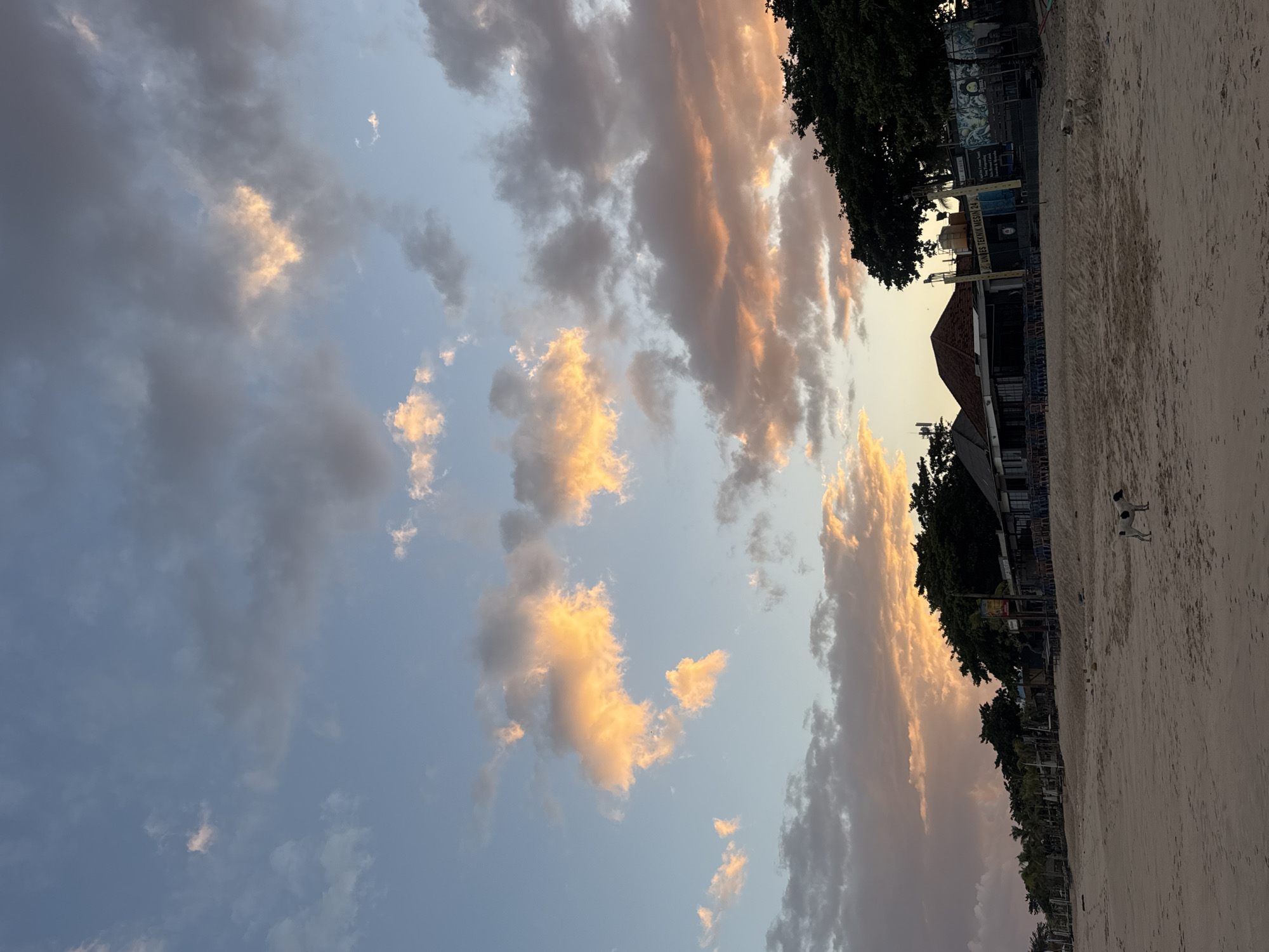 The image captures a tranquil beach scene in Kuta, Indonesia, during what appears to be late afternoon or early evening. The sky is a canvas of soft blue, transitioning to hues of orange and pink where clouds are catching the fading sunlight. These dramatic clouds, some dark and others illuminated with warm tones, create a striking contrast against the clear sky.
In the foreground, a sandy beach stretches towards the sea, showing subtle footprints indicating recent activity. A lone black and white dog stands alert on the sand, facing right. Further back, a row of low-lying buildings lines the beachfront, suggesting a commercial area with restaurants or shops. These structures have simple architectural designs, with some featuring red-tiled roofs and outdoor seating areas with numerous chairs and tables. Dense green trees are interspersed along the property line, adding a natural element to the developed area. The overall impression is one of a peaceful coastal setting, likely a popular spot for relaxation and dining, as evidenced by the numerous outdoor chairs.