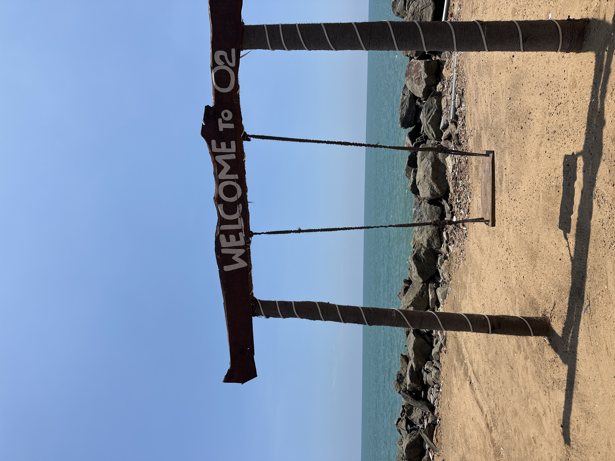 A swing set, labeled "WELCOME TO O2," is positioned at the edge of a sandy area adjacent to a rocky shore, with a view of the ocean under a clear sky. The location is Unknown, Unknown.