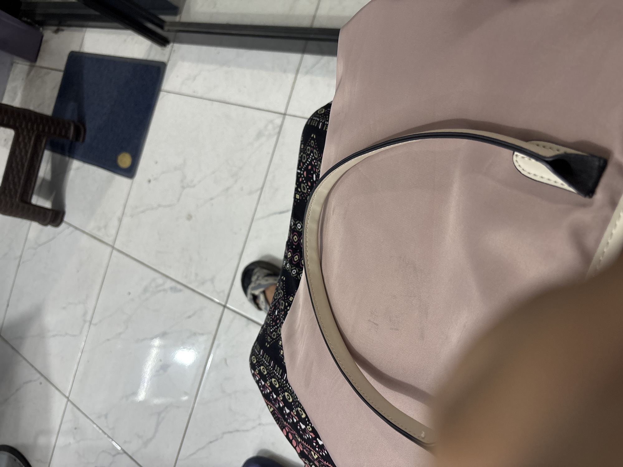 A person is seated on a tiled floor, with their legs crossed. They are wearing a patterned skirt and sandals. A pink handbag is in the foreground, with a strap visible. In the background, a dark brown plastic chair and a blue mat are visible on the tiled floor. The overall scene suggests an indoor environment, possibly a waiting area or a room in a residential property in Wanajaya, Indonesia.