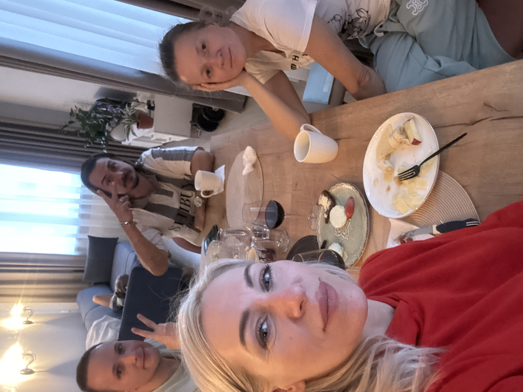 The image captures a social gathering around a wooden table laden with remnants of a meal - a partially eaten cake on a white plate with a fork, another decorative plate with food items, wine glasses (some empty, some filled), and mugs. Four individuals are visible at the table. A woman in a red sweater is taking a selfie. Another woman, wearing a white t-shirt, rests her head on her hand, looking towards the camera. A man, seated further away, holds up a peace sign. A fourth person, partially visible at the bottom left, also flashes a peace sign. The setting appears to be indoors, possibly a dining room or living area, evidenced by the presence of a sofa, curtains, and indoor plants. Based on the information provided in the prompt, the location is in the city of Unknown, Russia.