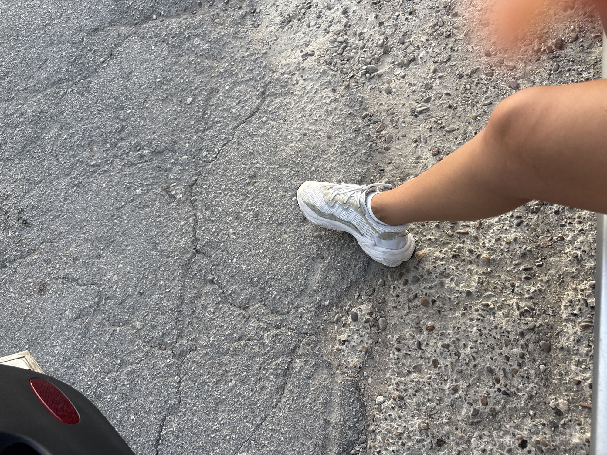 The image shows a person's right leg and foot wearing a white athletic shoe, standing on a cracked asphalt surface. A black object with a red reflective element is partially visible in the upper left corner, likely part of a vehicle. The ground appears to be a road or pathway in Cimișlia, Moldova, characterized by its rough texture and visible cracks. The lighting suggests it is daytime.