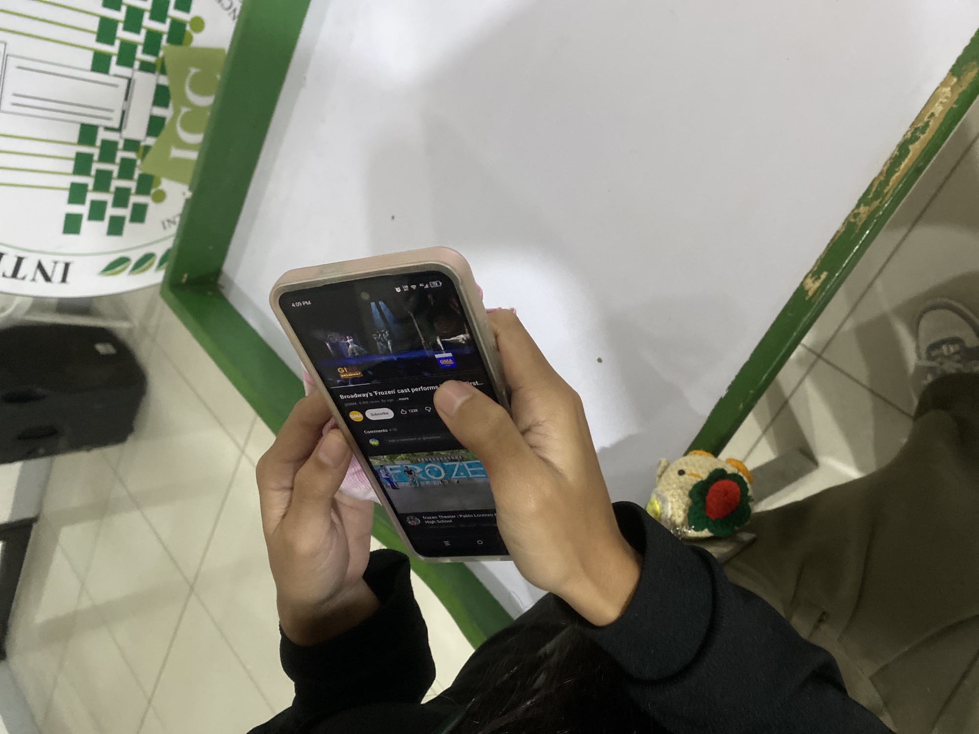 The image captures a top-down view of a person holding a smartphone, likely indoors in Davao City, Philippines. The subject's hands, with manicured nails, cradle a light-colored smartphone with a visible screen displaying what appears to be a YouTube-like interface. The time on the phone screen reads "4:09 PM".
The primary activity is the viewing of a video titled "Broadway's 'Frozen' cast performs first..." from the channel "GMA," which has garnered "8.3M views." Below the video player, options for "Subscribe," "122K" likes, and "Comments 41K" are visible. A suggested video thumbnail features "FROZE" and is titled "Frozen Theater : Pablo Lorenzo High School."
The setting suggests an institutional environment. To the left, part of a white surface with a green logo is visible, featuring the text "INTERNATIONAL COLLEGES" and "ICC". A distinct green frame outlines the white surface, with some wear and peeling paint visible on its edge. On the right, next to the person's right hand, sits a small, light-colored crocheted item resembling a chick or bird, adorned with red and green elements. The floor is tiled in light squares, and parts of what appear to be the person's dark clothing and footwear are also visible.